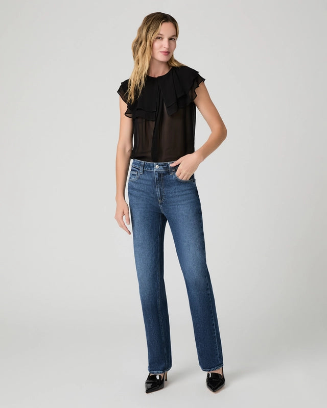 A woman stands against a plain background, wearing a sheer black ruffled top, the Paige Stevie High Rise Straight Leg - Jetty jeans in a medium-blue wash, and black pointed-toe shoes.
