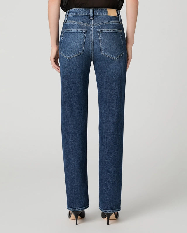 A person is seen from the back wearing the Paige Stevie High Rise Straight Leg - Jetty jeans in a medium-blue, heritage-inspired wash, paired with a black top and black pointed-toe shoes against a plain light background.