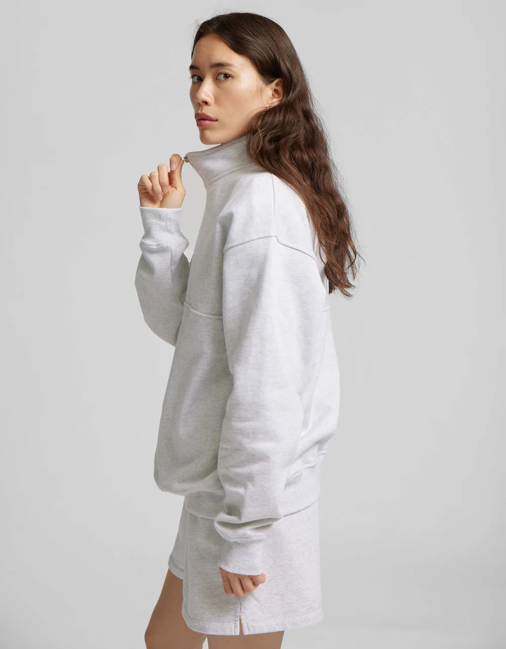 A person with long brown hair wears a gray Organic Quarter Zip from Colorful Standard and shorts, glancing over their left shoulder against a plain backdrop.