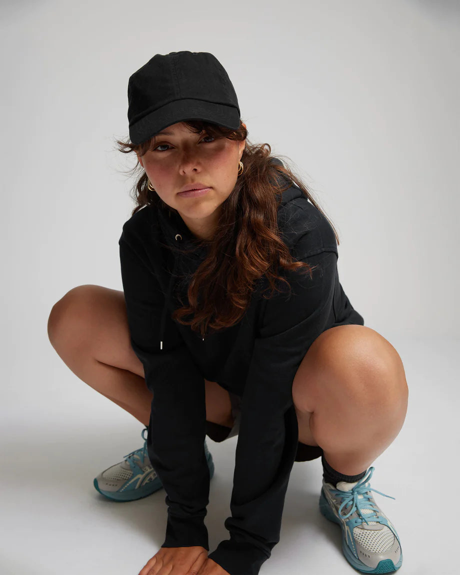 A woman crouching down in front of a white background, showcasing the soft and comfortable feel of Colorful Standard's Organic Cotton Cap, free from harmful chemicals.