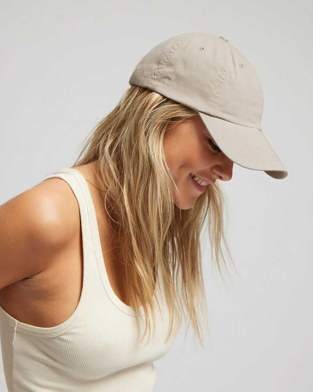 Woman smiling downward wearing a beige baseball cap and a soft and comfortable Colorful Standard organic cotton tank top.