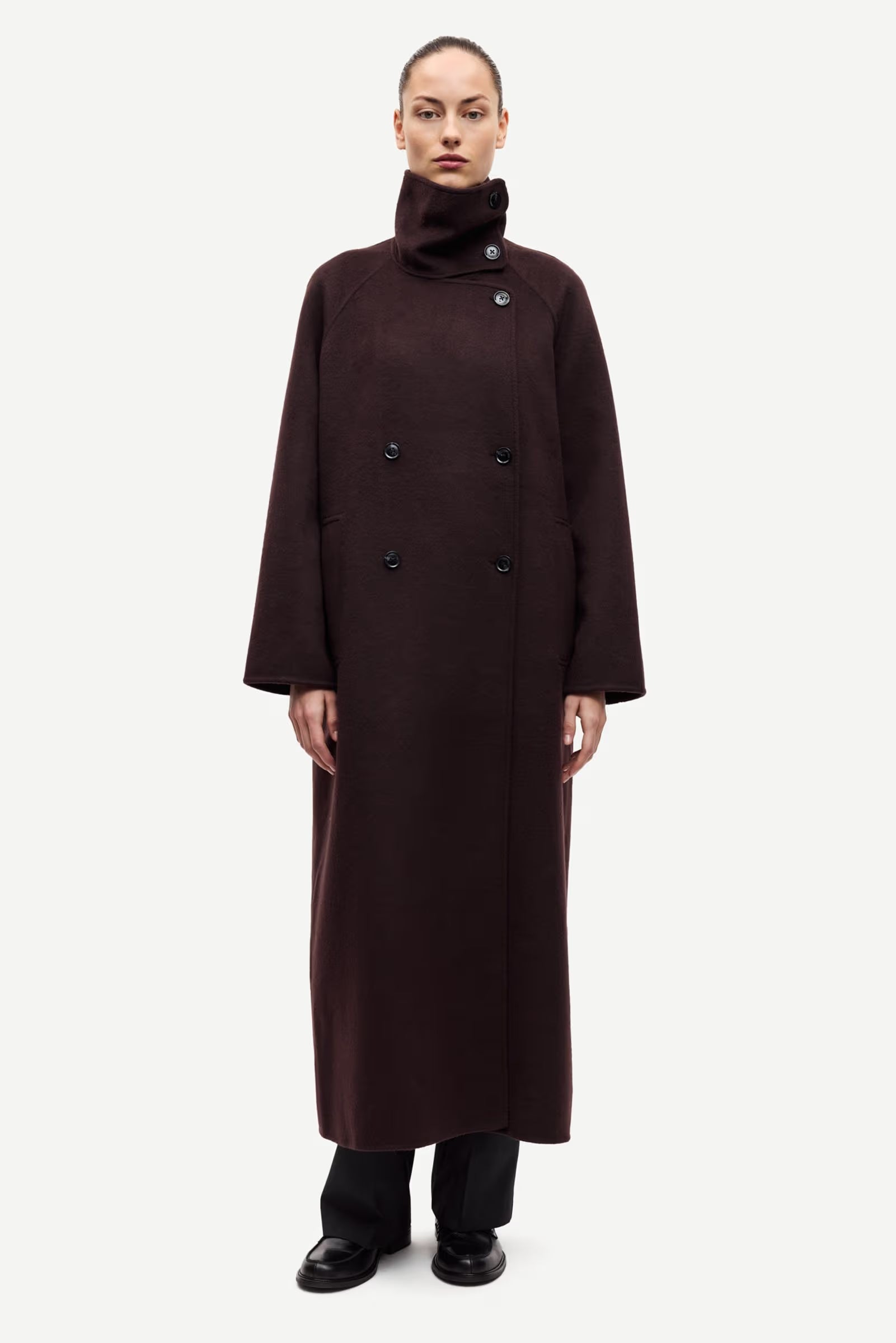 A person wearing the Sahanne Coat in Black Coffee by Samsøe Samsøe—a long, double-breasted felted coat with a high collar and oversized fit—stands against a plain white background.