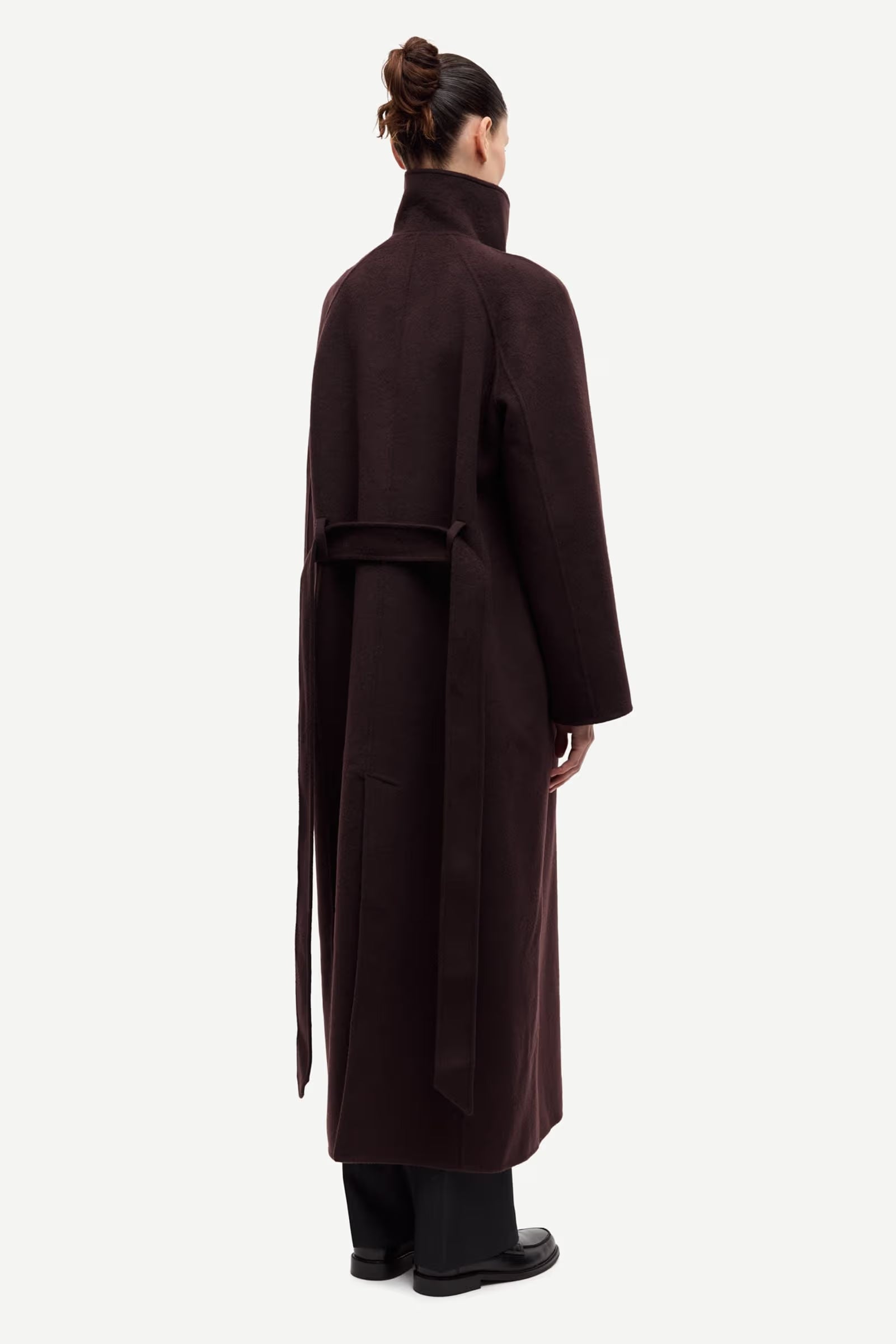 A person with their hair in a bun is seen from the back wearing the Samsøe Samsøe Sahanne Coat in Black Coffee—a long, oversized felted coat with a high collar and belt—set against a plain background.