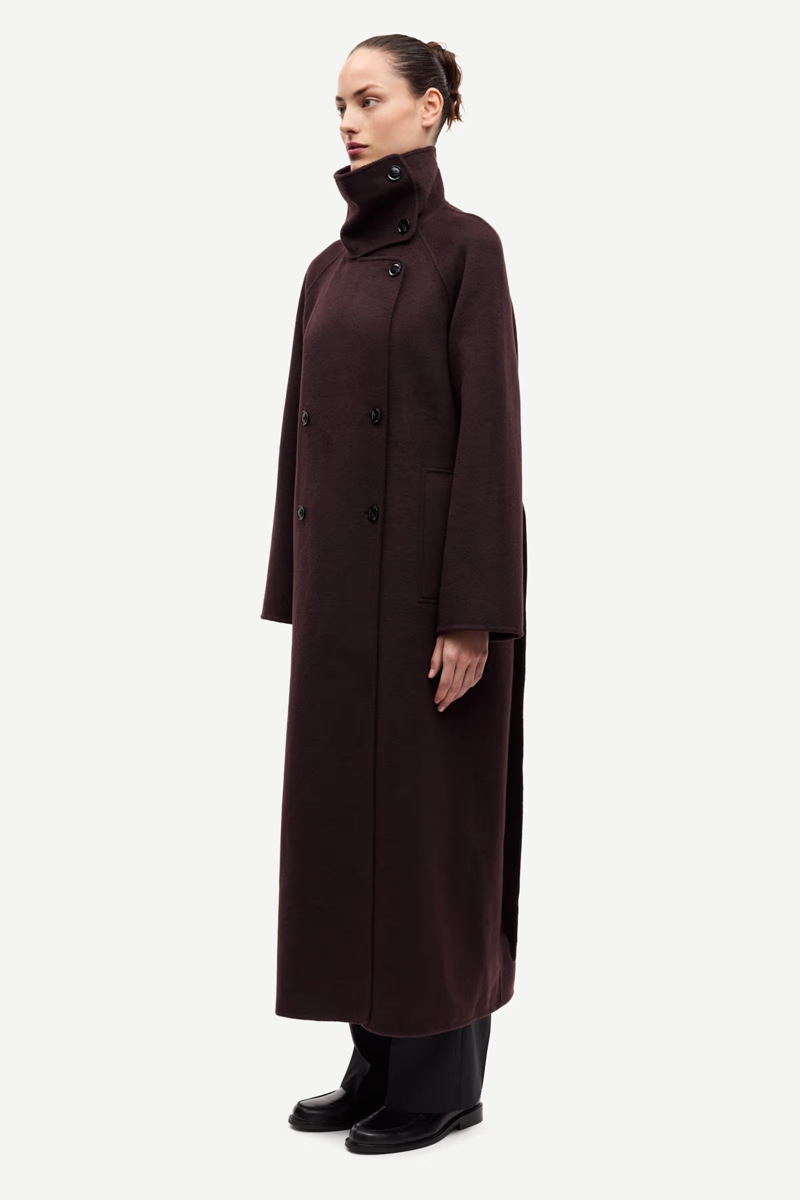 A woman stands in profile against a plain light background, wearing the Samsøe Samsøe Sahanne Coat in Black Coffee—a long, dark brown double-breasted felted coat with a high collar—paired with black shoes.