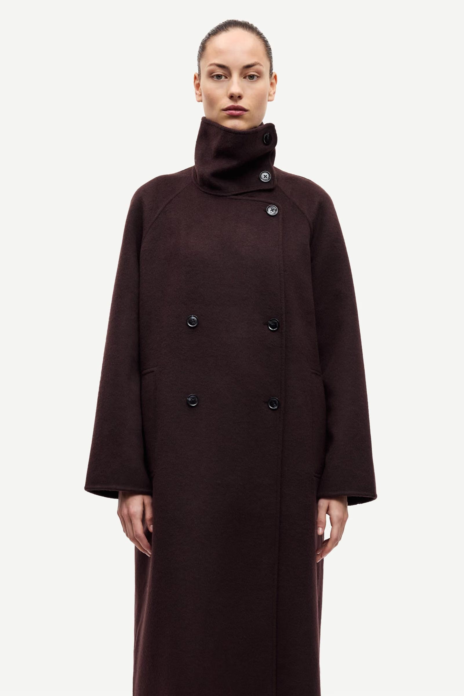 A woman wearing the Samsøe Samsøe Sahanne Coat in Black Coffee—a long, double-breasted felted coat with a high collar, large buttons, and an oversized fit—stands against a plain white background.