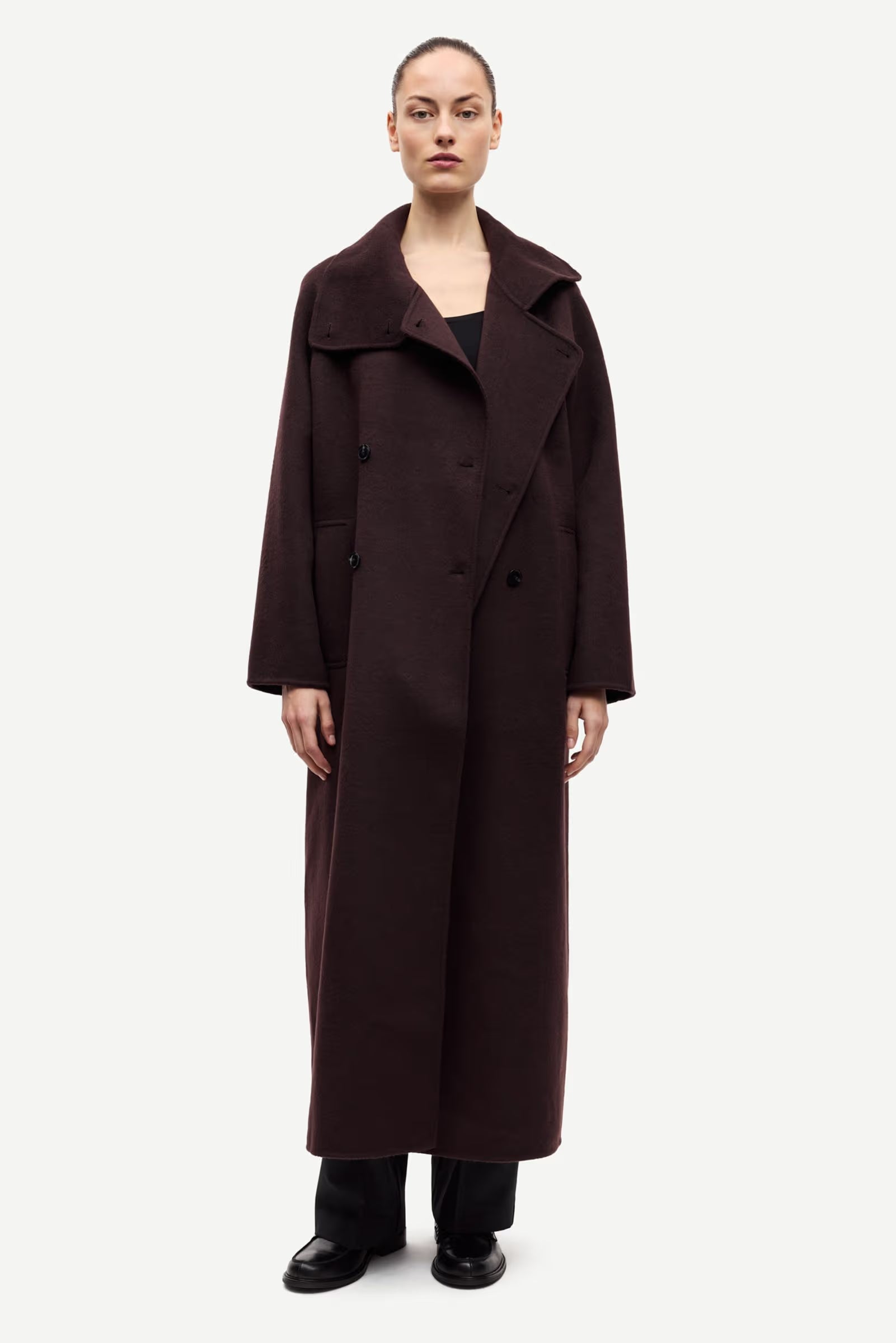 A person stands facing forward, wearing the Samsøe Samsøe Sahanne Coat in Black Coffee with an oversized fit, paired with black pants and black shoes against a plain white background.