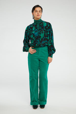 person in a studio wears green cords and a green/black striped polo neck under the green floral Rese blouse by Fabienne Chapot