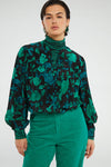person in a studio wears green cords and a green/black striped polo neck under the green floral Rese blouse by Fabienne Chapot