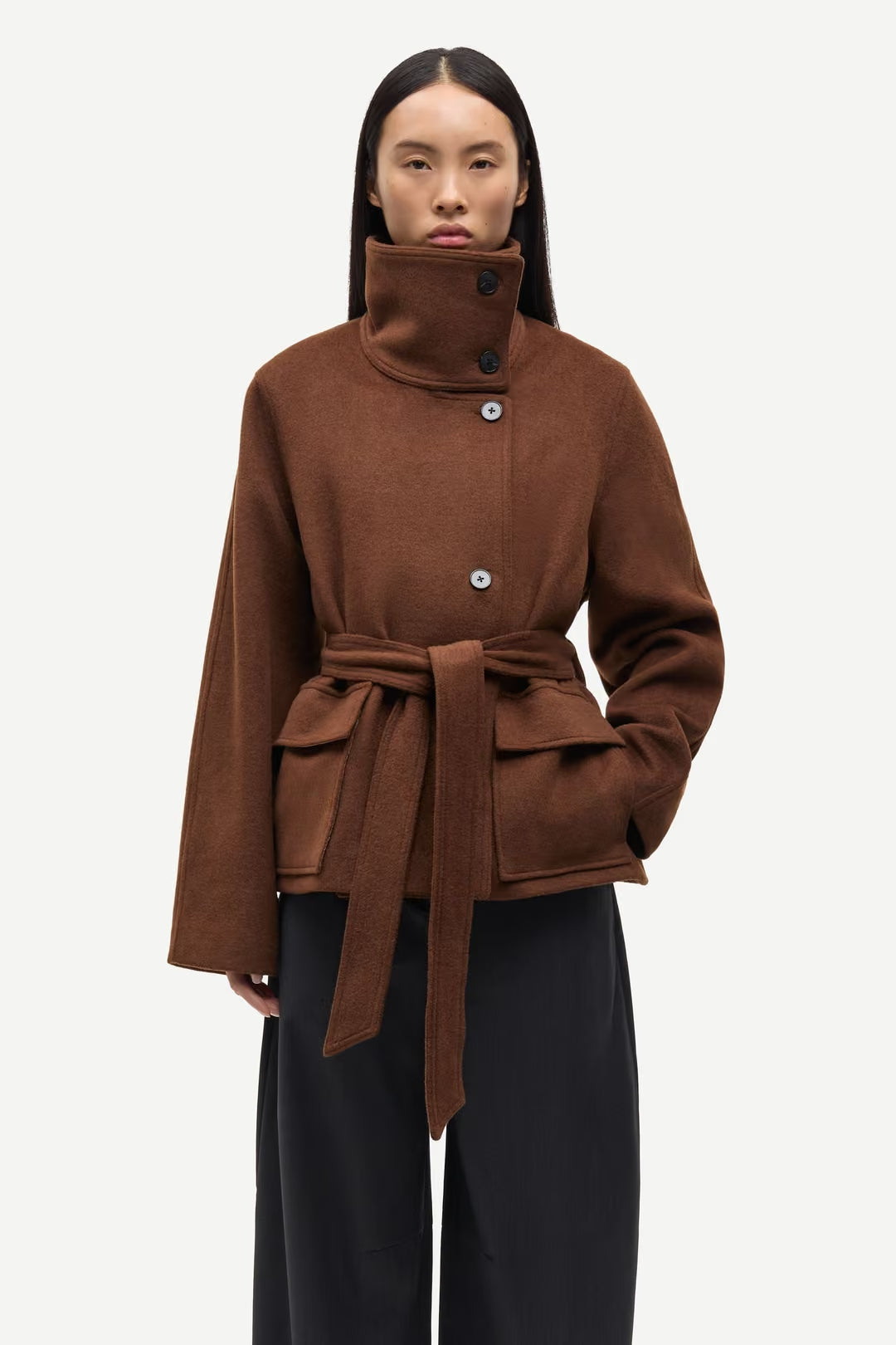 A person wearing the Sagia Jacket in brown by Samsøe Samsøe, featuring a high collar and wide sleeves, paired with loose black pants, stands against a plain white background.
