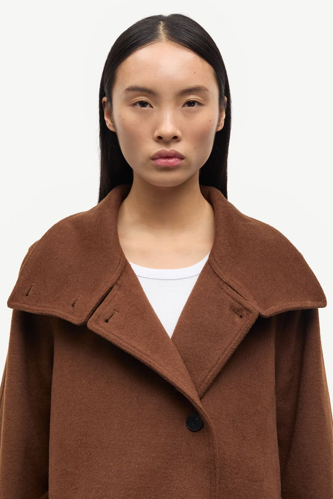 A person with straight dark hair faces forward against a plain light background, wearing the Sagia Jacket in brown by Samsøe Samsøe over a white top.