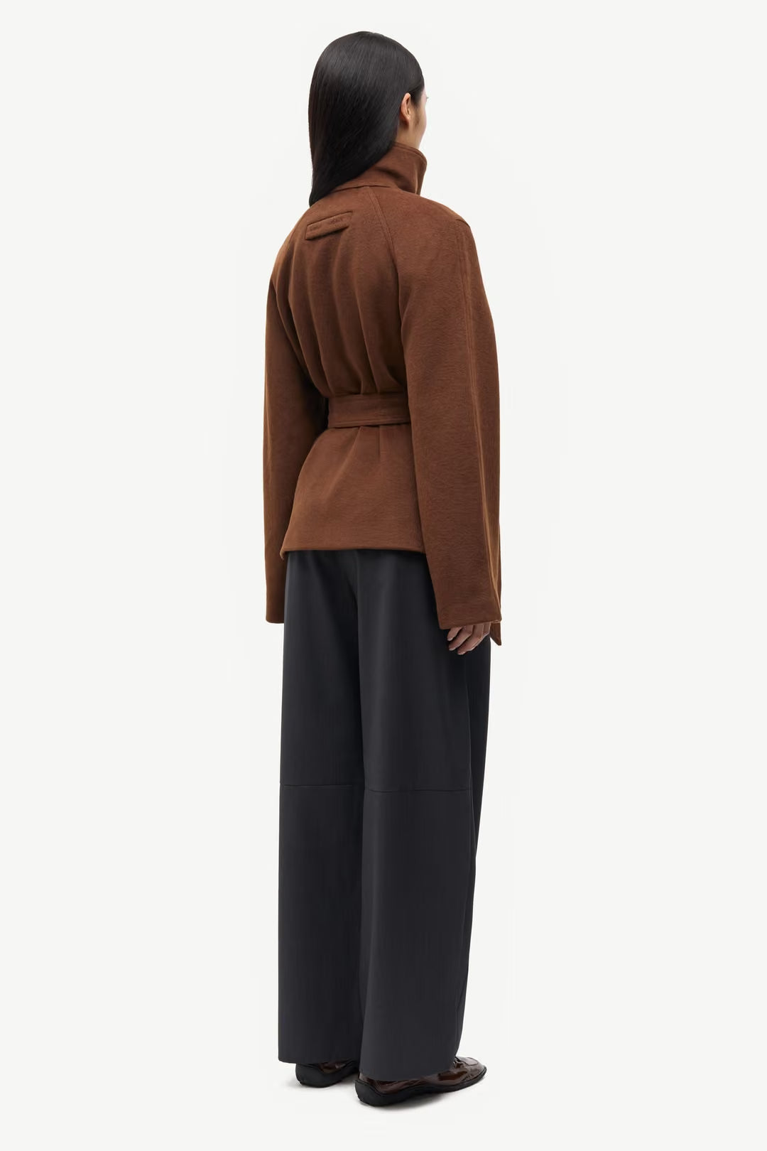A person stands facing away, wearing the Samsøe Samsøe Sagia Jacket in brown, featuring a tie belt, paired with wide-leg black pants. Their straight dark hair is visible from the back against a plain white background.