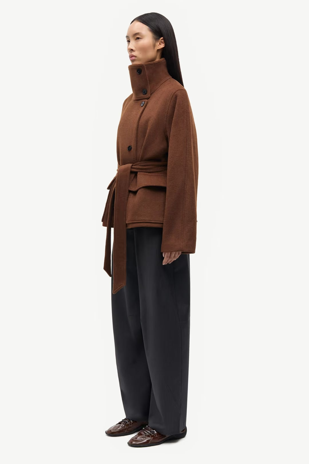 A woman stands in profile against a plain white background, wearing the Sagia Jacket in brown by Samsøe Samsøe, styled with wide-leg dark trousers and dark shoes.