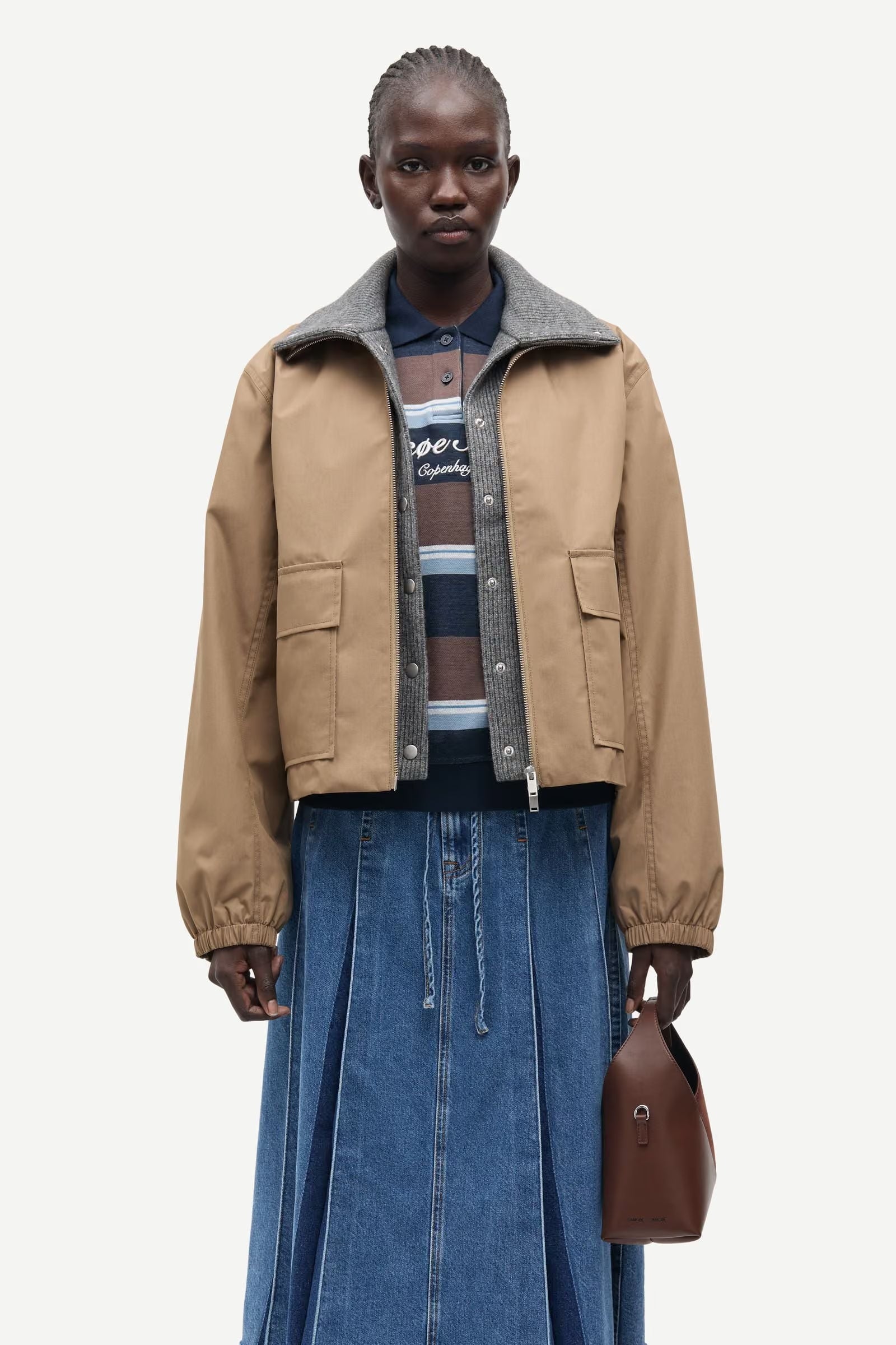 Wearing the Samsøe Samsøe Sabianco Jacket in lead grey over a striped shirt and blue denim skirt, the individual holds a brown handbag while standing against a plain white background.