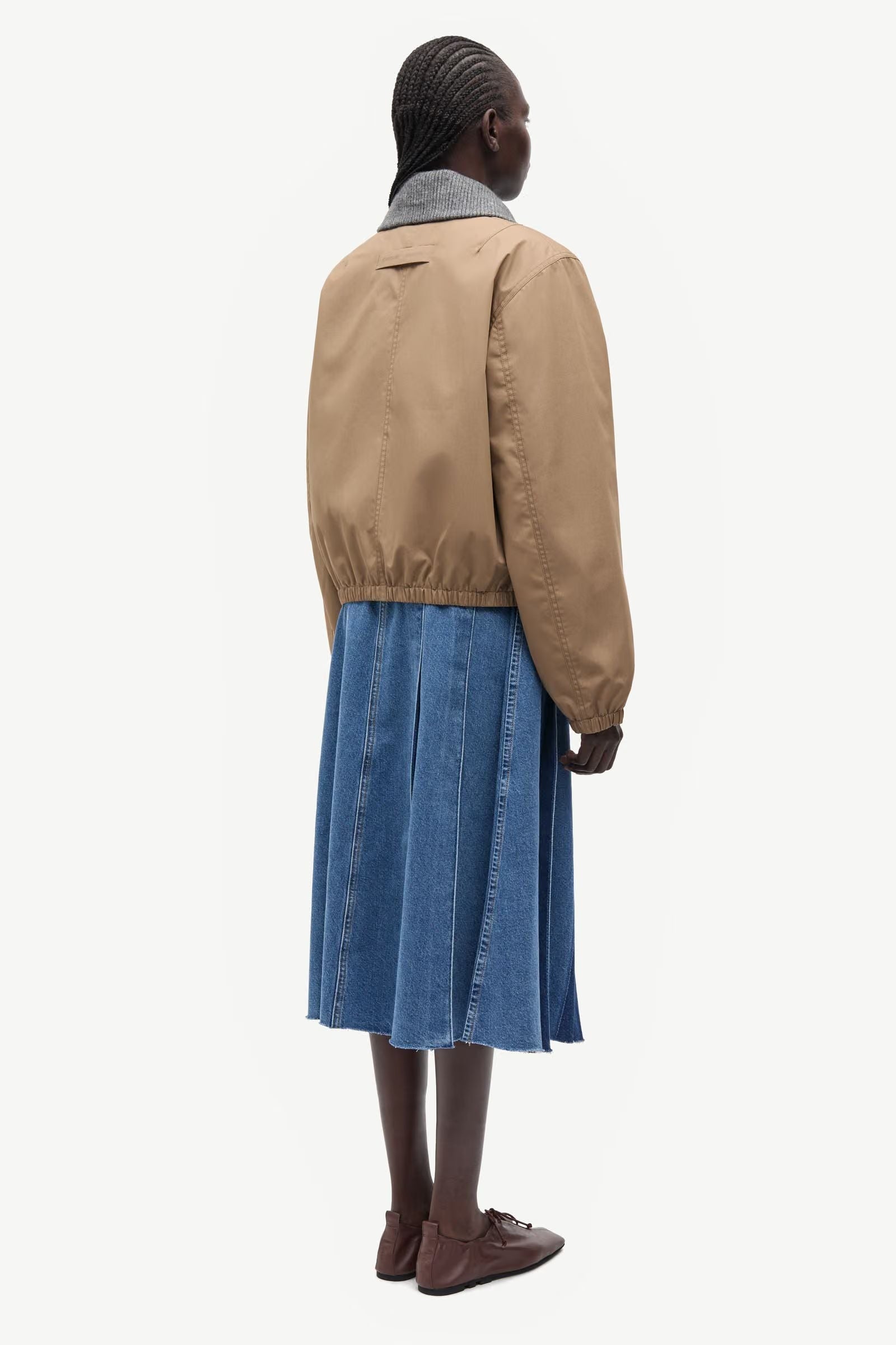 A person wearing the Sabianco Jacket in Lead Grey by Samsøe Samsøe stands facing away against a white background, paired with a blue pleated denim skirt and brown shoes.