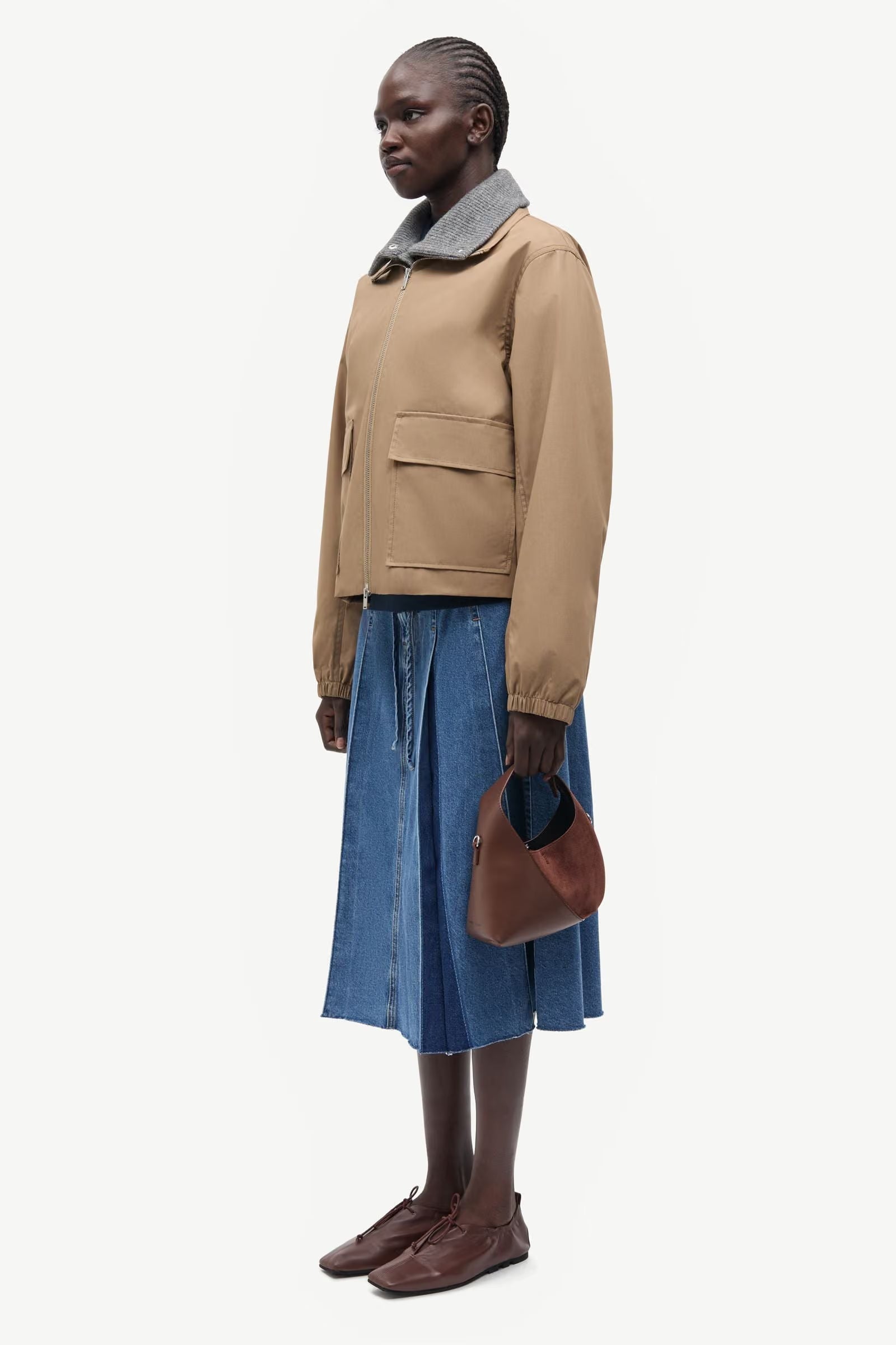 A woman stands against a plain white background wearing the Samsøe Samsøe Sabianco Jacket in lead grey, paired with a blue denim skirt, brown shoes, and holding a small brown purse.