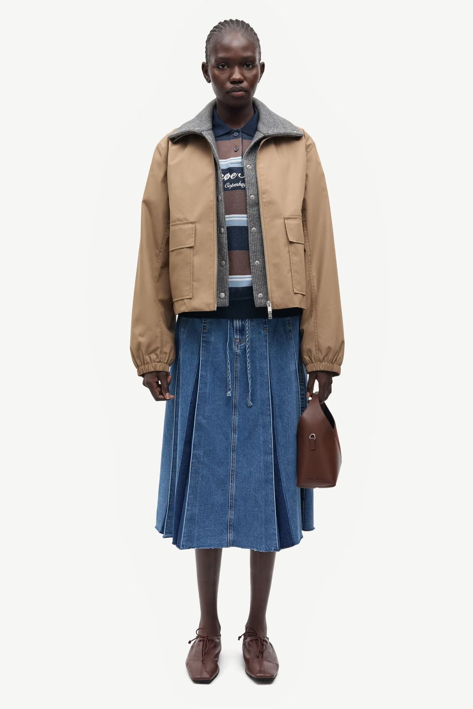 A person stands facing forward against a white background, wearing the Samsøe Samsøe Sabianco Jacket in lead grey, paired with a striped sweater, blue pleated denim skirt, brown shoes, and holding a small brown handbag.