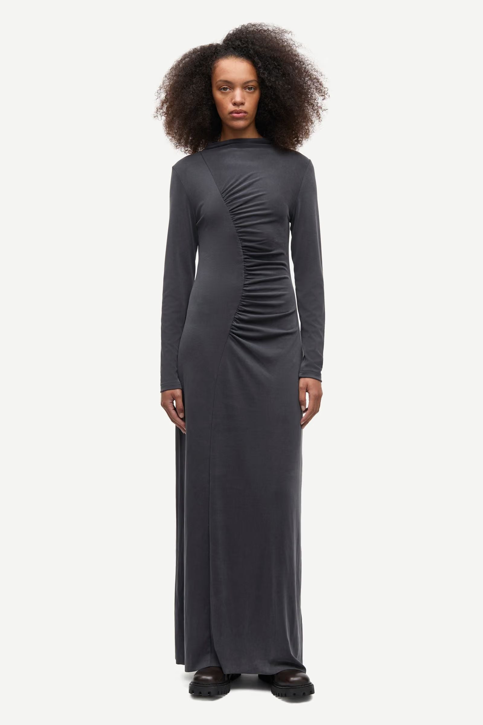 A person wearing the Samsøe Samsøe Saandressa Is dress in black—a long-sleeved, floor-length jersey maxi dress with center ruching detail—stands against a plain white background.