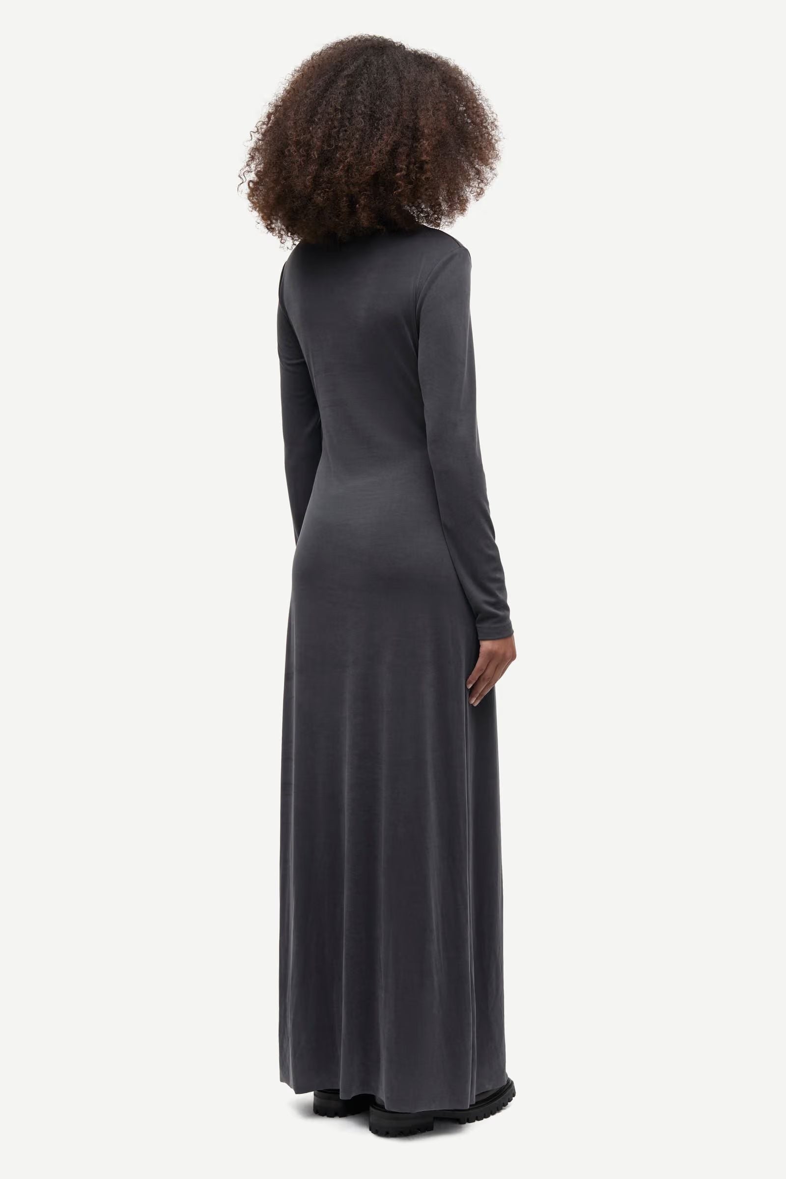 A person stands facing away, wearing the Saandressa Is dress in black by Samsøe Samsøe. The long-sleeved jersey maxi dress features soft fabric with ruched detailing and drapes elegantly to the floor against a plain white background.
