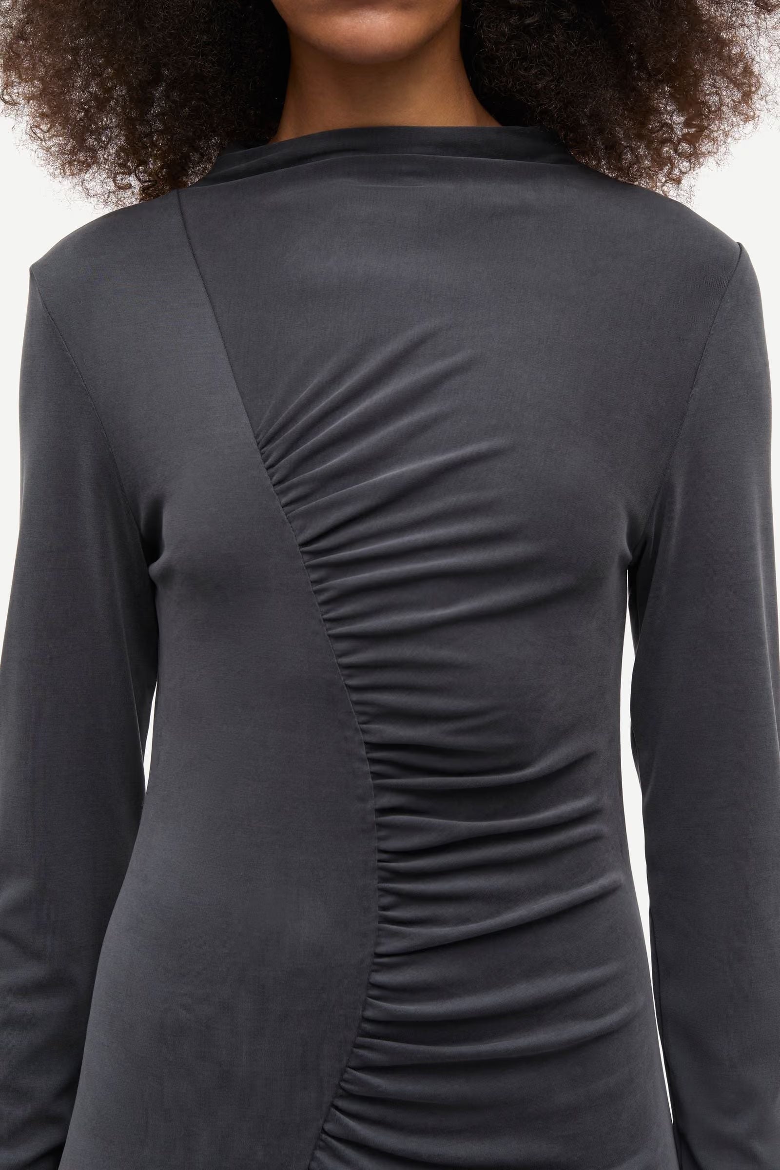 A model wears the Samsøe Samsøe Saandressa Is dress in black, a long-sleeved style with a high neckline and asymmetrical ruched detailing across the front.