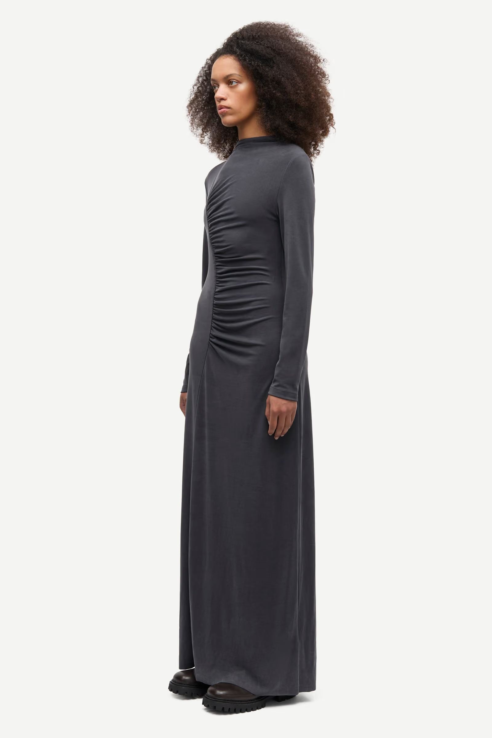 A woman stands against a plain white background wearing the Saandressa Is dress in black by Samsøe Samsøe, a long-sleeve jersey maxi dress featuring ruched waist detailing, paired with black shoes.