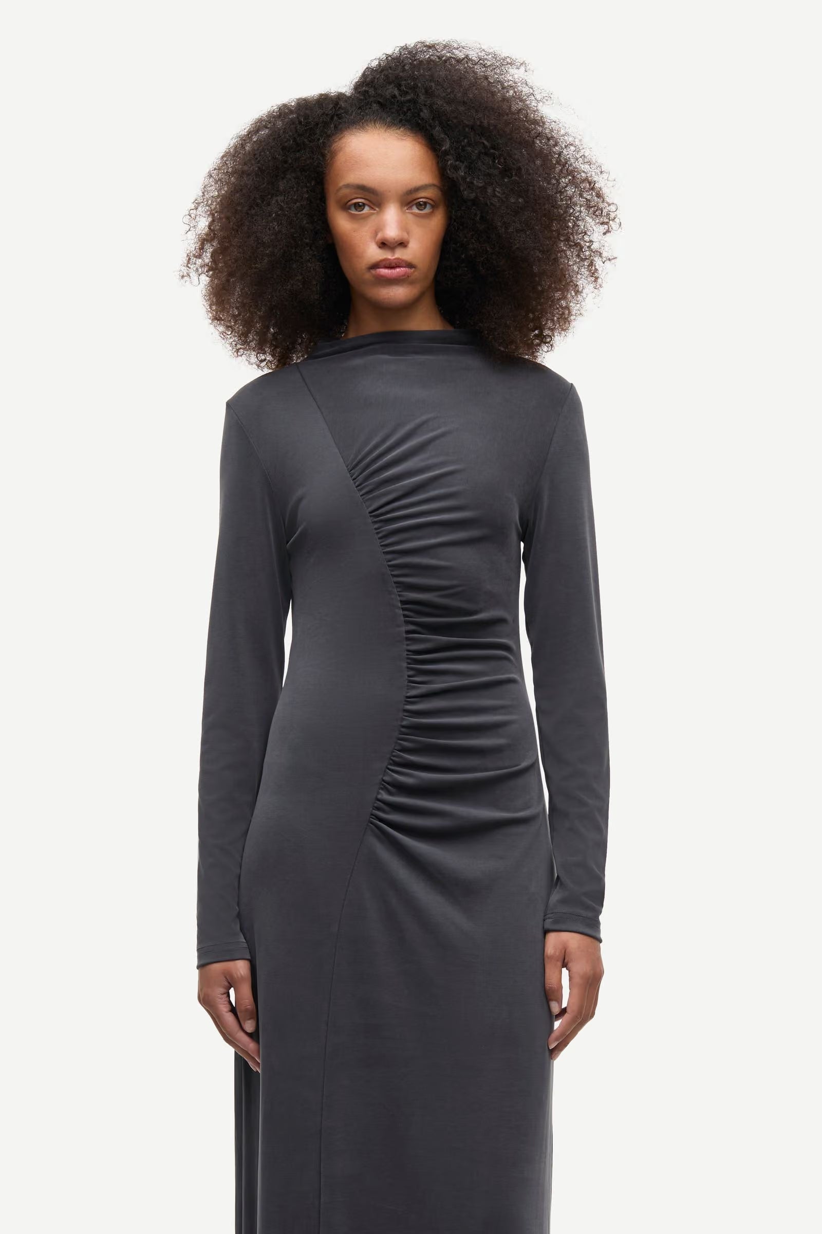 A person wearing the Samsøe Samsøe Saandressa Is dress in black—a long-sleeved jersey maxi dress featuring asymmetrical ruched detailing on the front—stands against a plain white background.