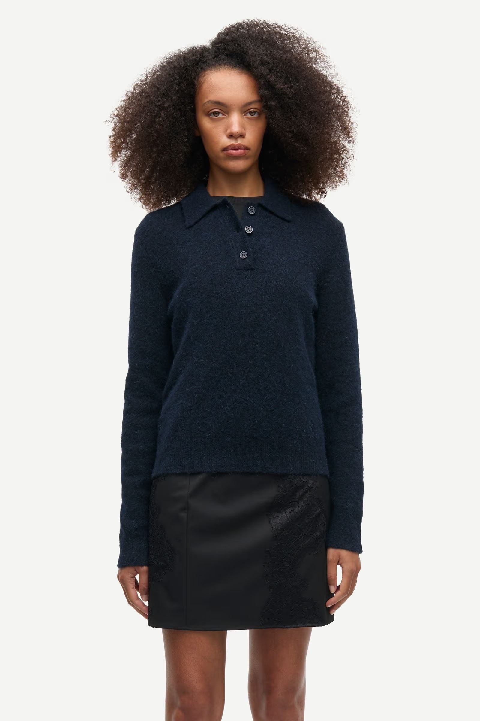 A woman stands facing forward against a plain white background, wearing the Samsøe Samsøe Sanoura LS Polo in navy—an elegant long-sleeve polo collar sweater with buttons, crafted from a soft blend of wool, alpaca, and recycled polyamide—paired with a black mini skirt.