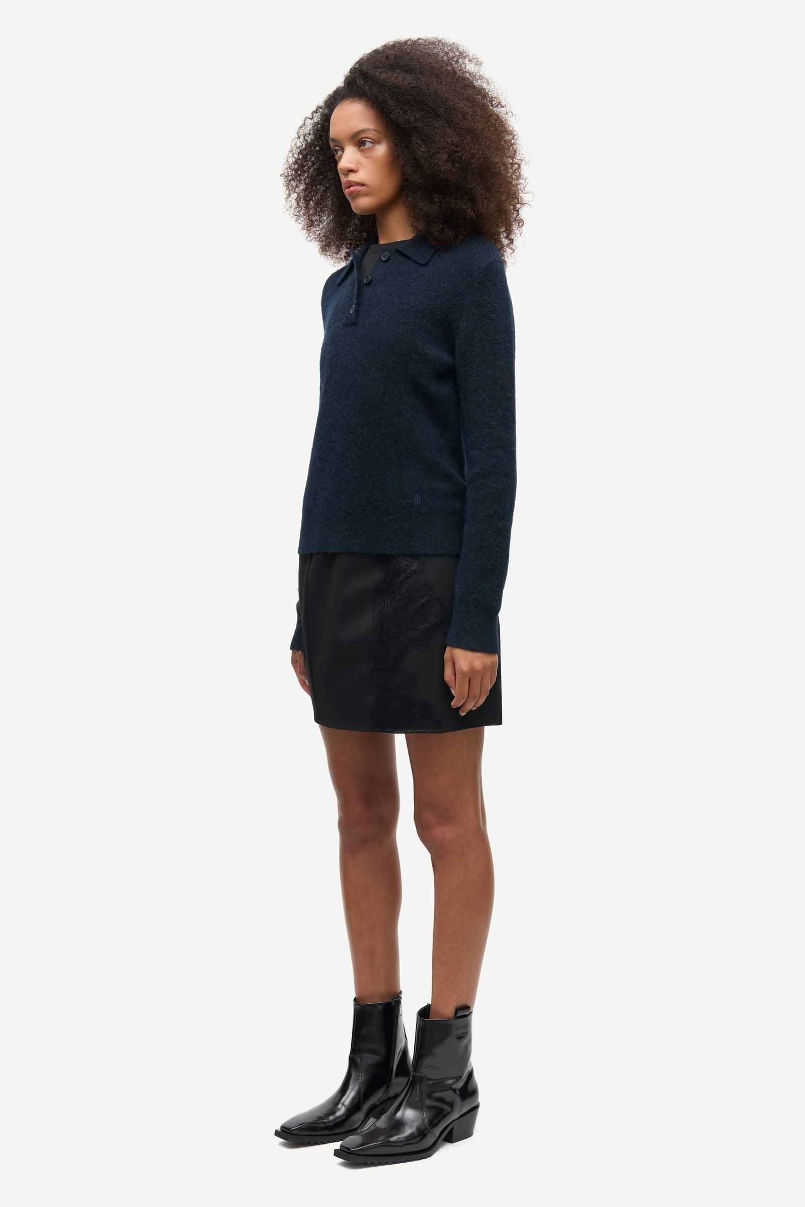 A person stands facing left, wearing the Sanoura LS Polo in navy by Samsøe Samsøe, paired with a black skirt and black ankle boots against a plain white background.