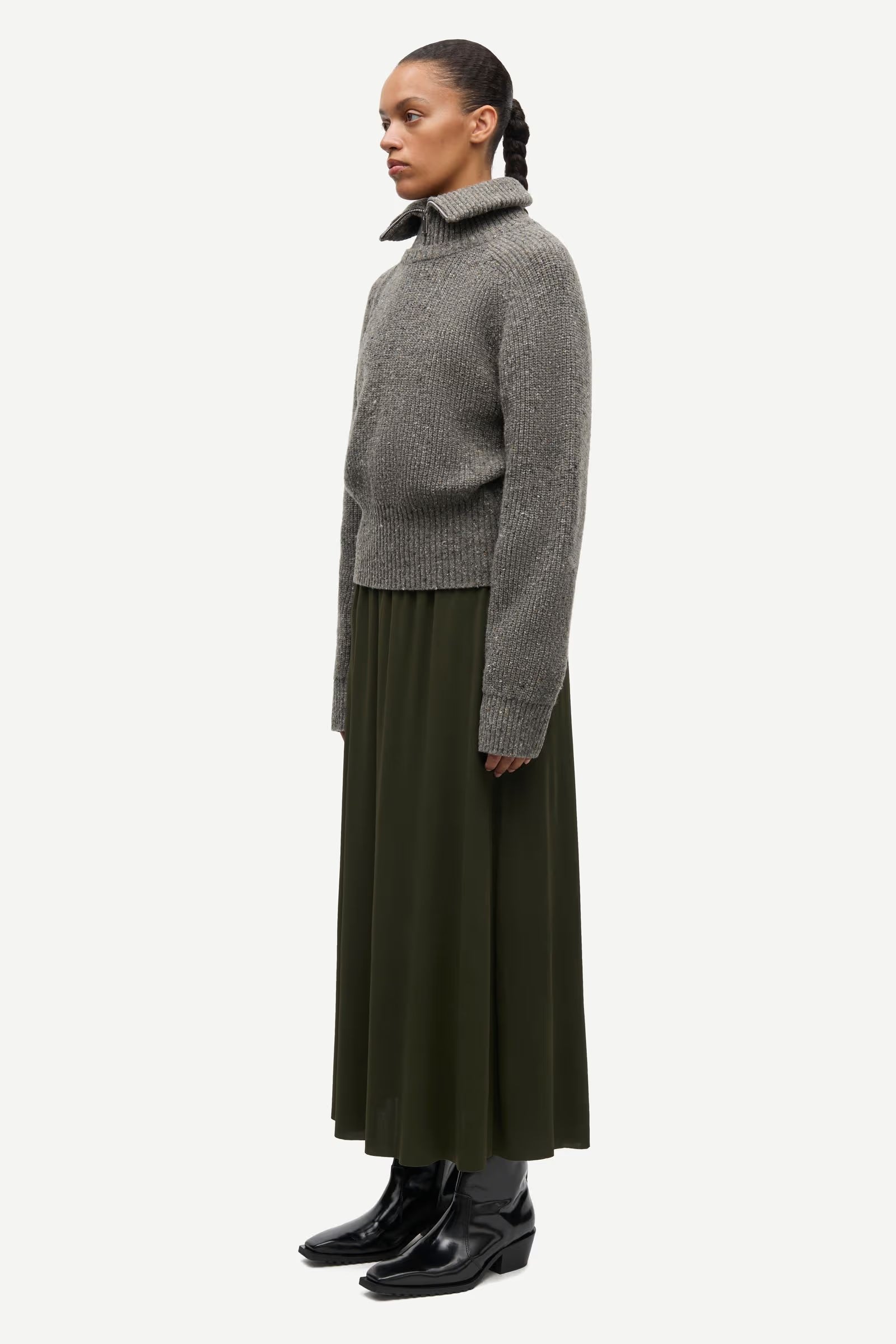 A person stands in profile against a plain white background, wearing the Samsøe Samsøe Saamandy Turtleneck in Lead Grey Melange, paired with a long olive green skirt and black ankle boots.