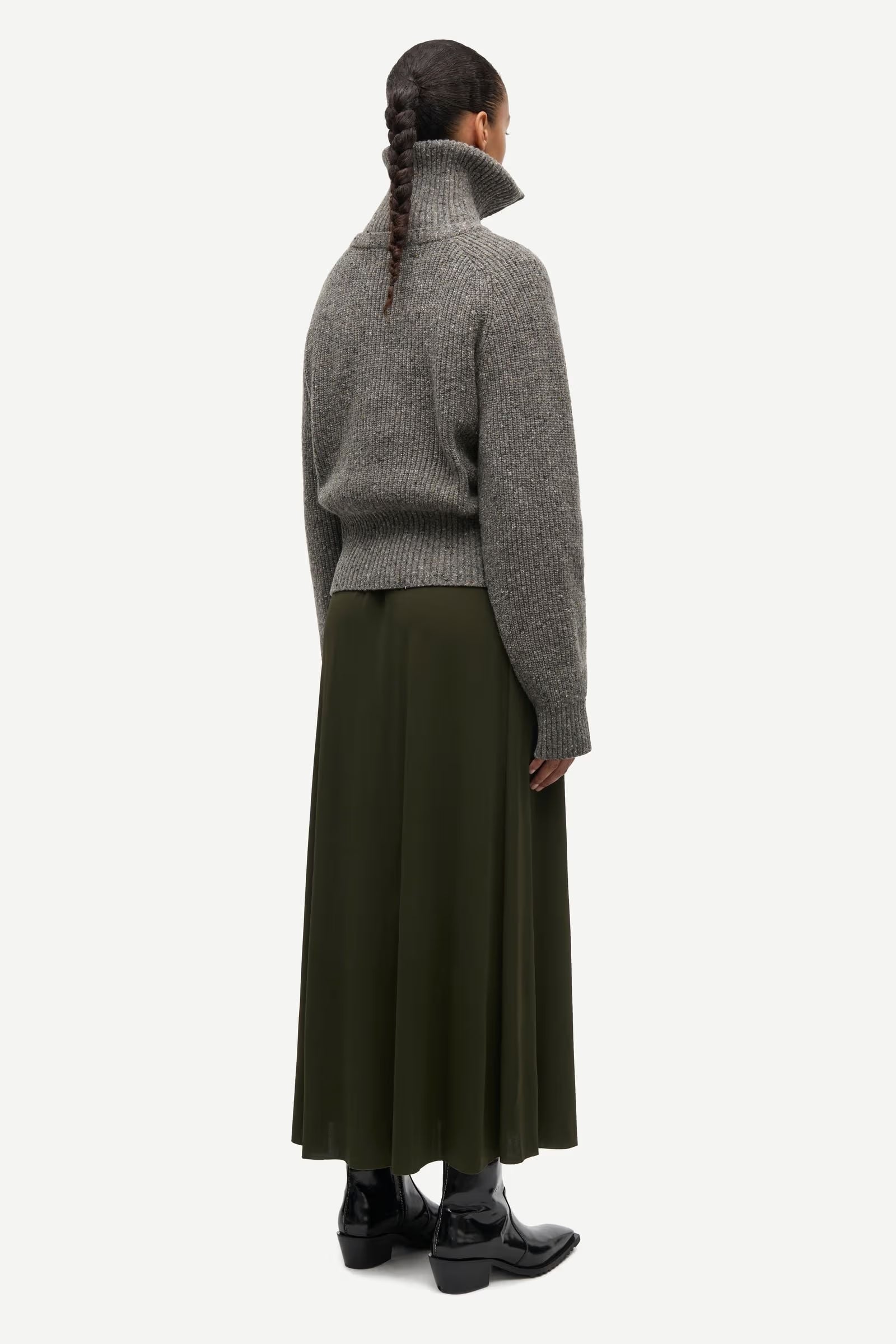 Facing away from the camera, the person wears the Samsøe Samsøe Saamandy Turtleneck in Lead Grey Melange as a turtleneck sweater, paired with an olive green midi skirt and black ankle boots. Their hair is styled in a braid against a plain white background.