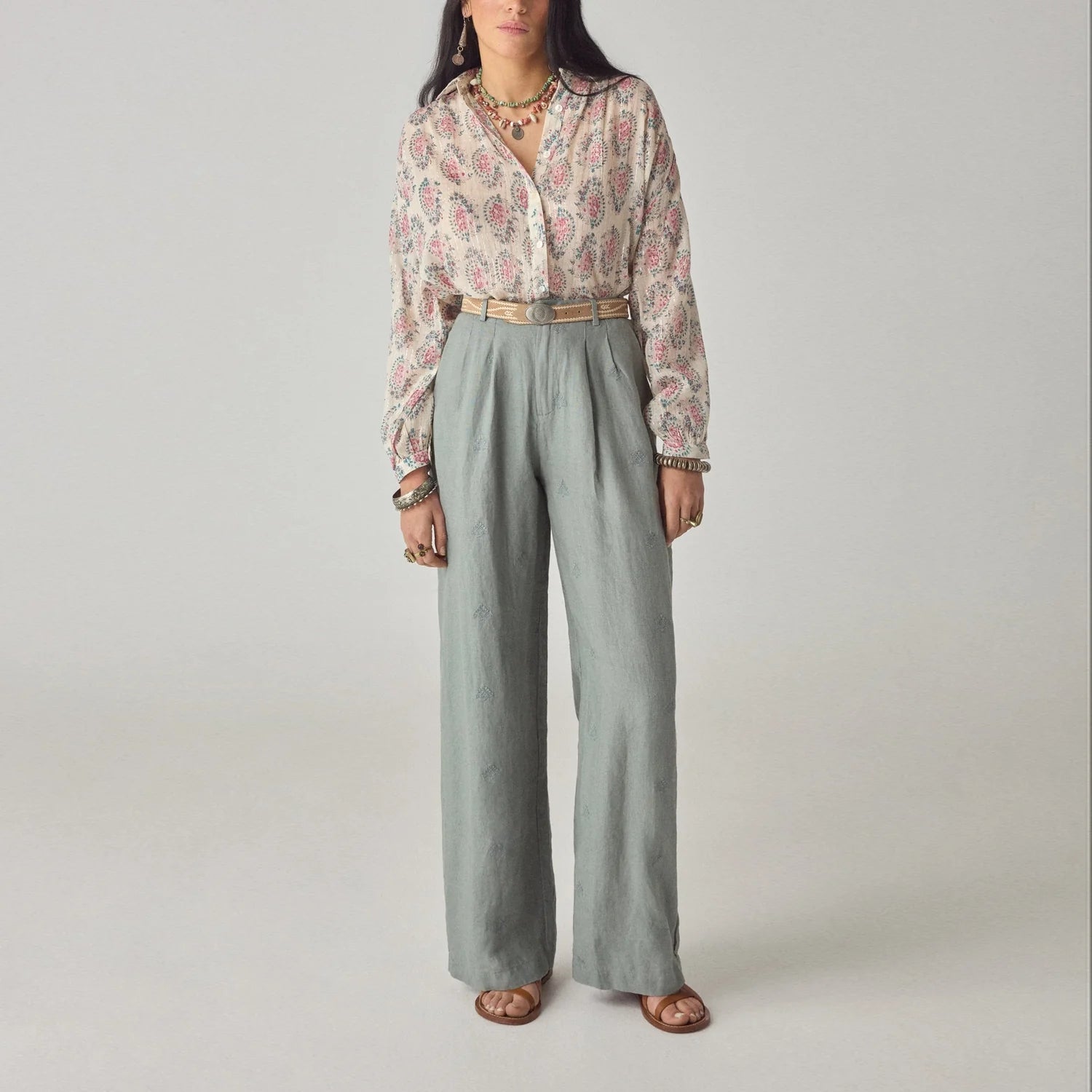 A woman wears the Mary Blouse - Ivory by Maison Hotel, featuring balloon sleeves, tucked into high-waisted gray trousers with a necklace, rings, and sandals against a plain background.