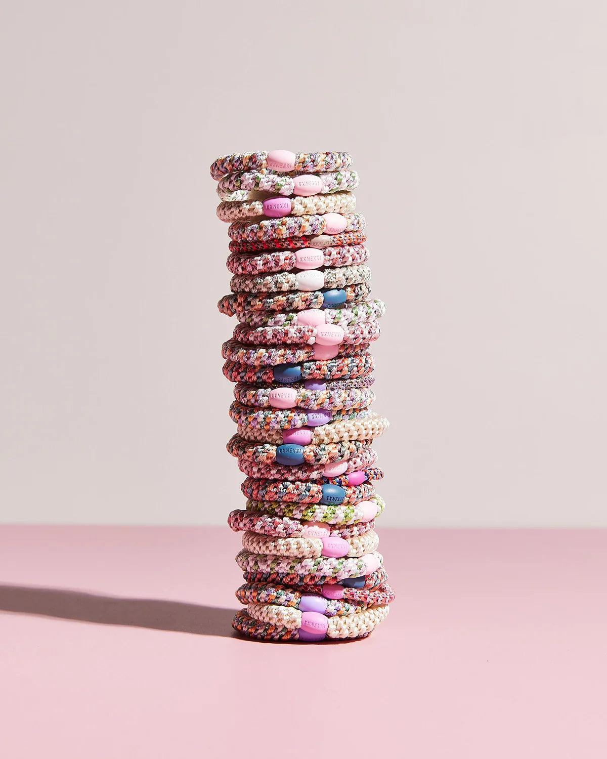 A tall stack of KKNEKKI Hair Tie - Mix, featuring colorful patterns and gentle grip with pink, purple, and white clasps, is arranged on a pink surface against a light background.