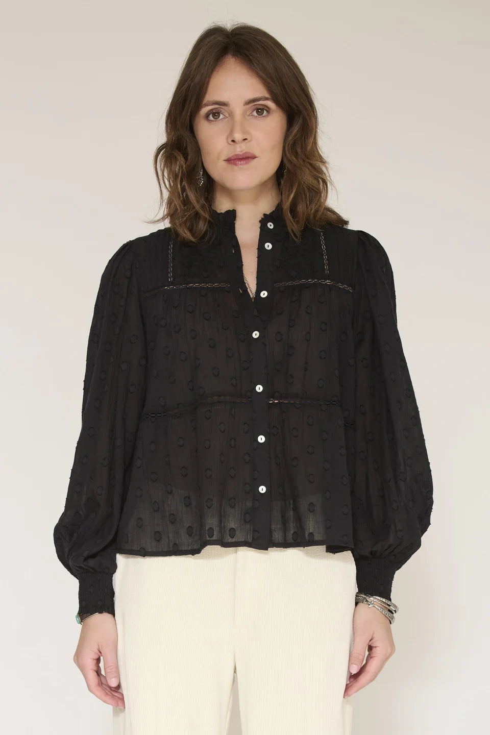 A woman modeling a black long-sleeve blouse with a subtle tonal pattern, button front, and decorative stitching details, paired with off-white trousers against a plain light background.
