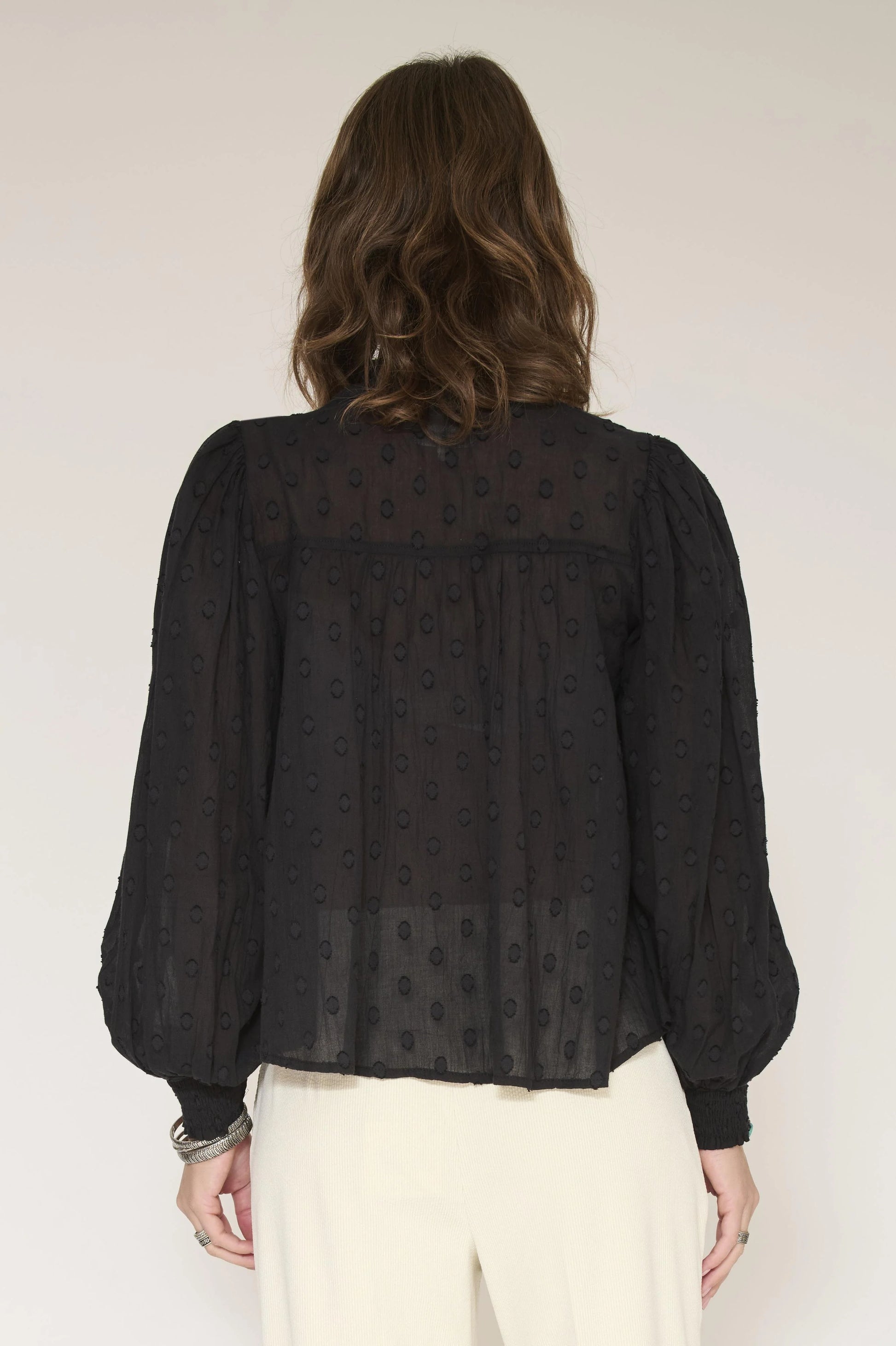 A woman viewed from the back wearing a black sheer long-sleeve blouse with a subtle allover dot pattern and cream-colored pants, set against a plain light beige background.