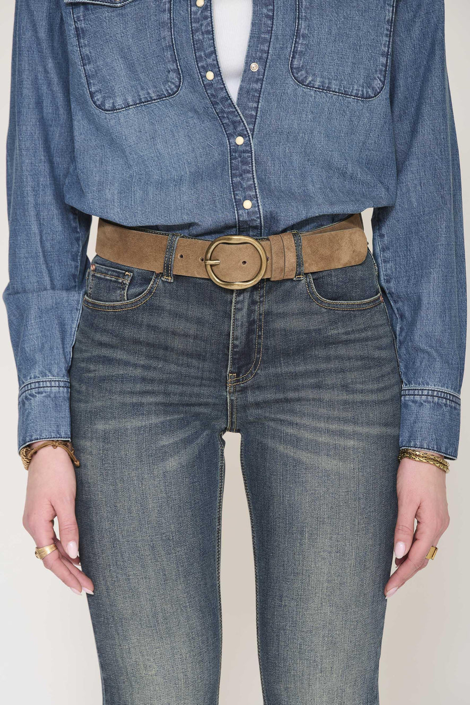A person is wearing a blue denim shirt, blue jeans, and the MKTStudio Duno Belt - Suede, a tan suede leather belt with a round silver buckle, standing with their hands by their sides.