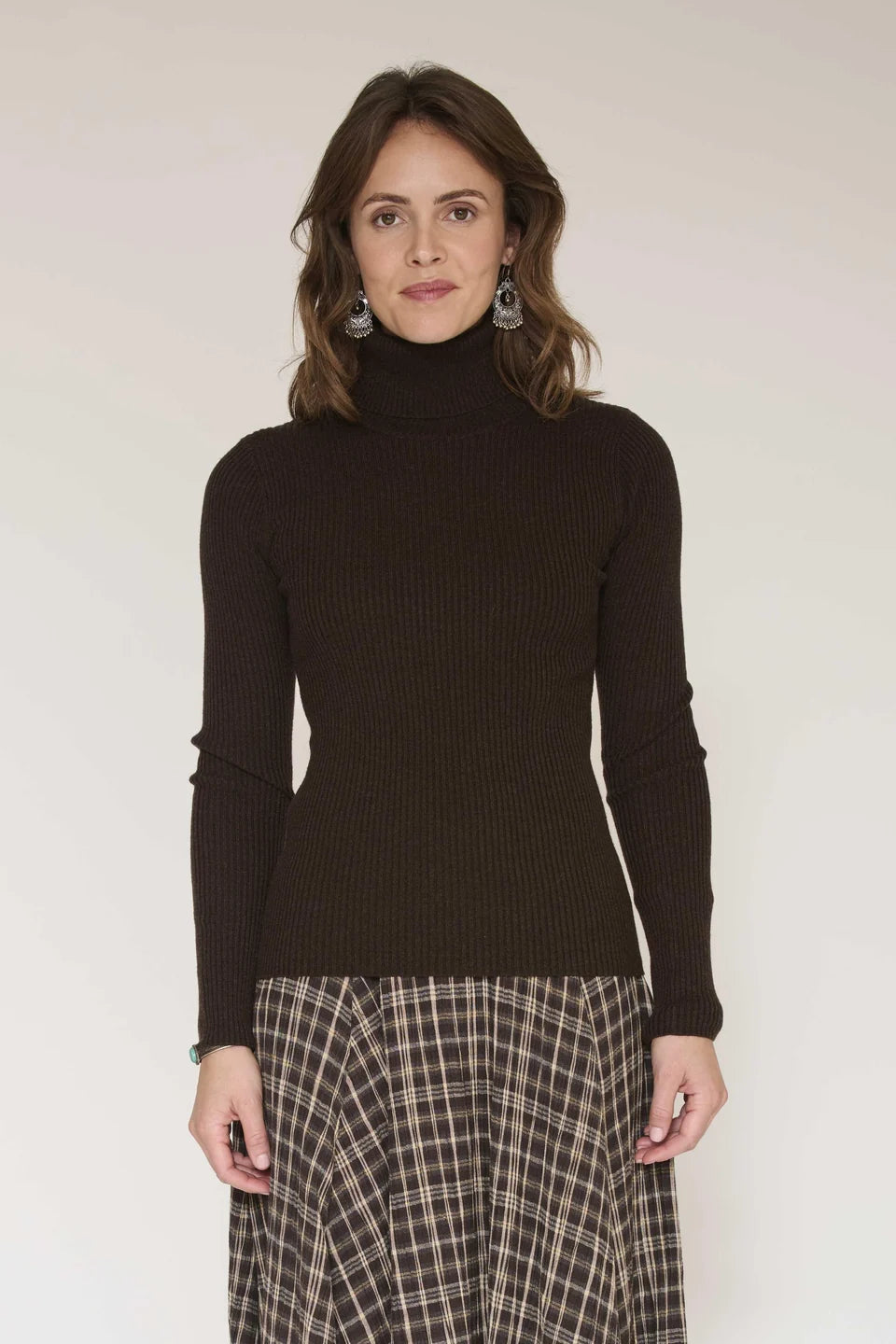 A woman wearing the Klark Sweater in brown by MKTStudio and a plaid skirt stands against a plain light background, facing forward with a neutral expression.