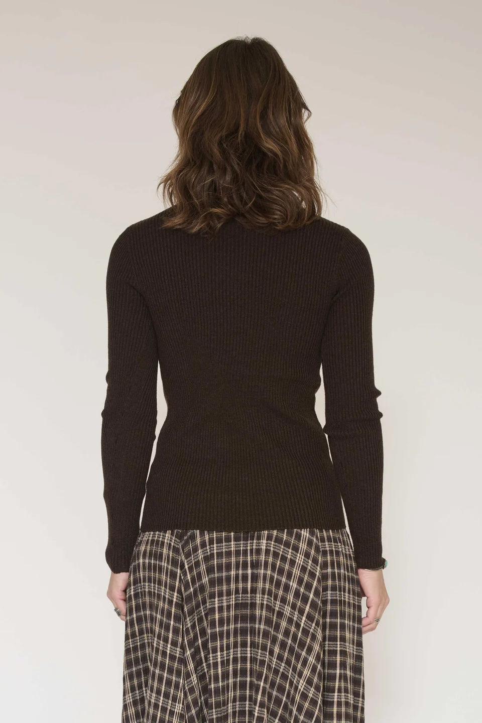 A person with shoulder-length brown hair, wearing the MKTStudio Klark Sweater in brown and a plaid skirt, is standing with their back to the camera against a plain background.