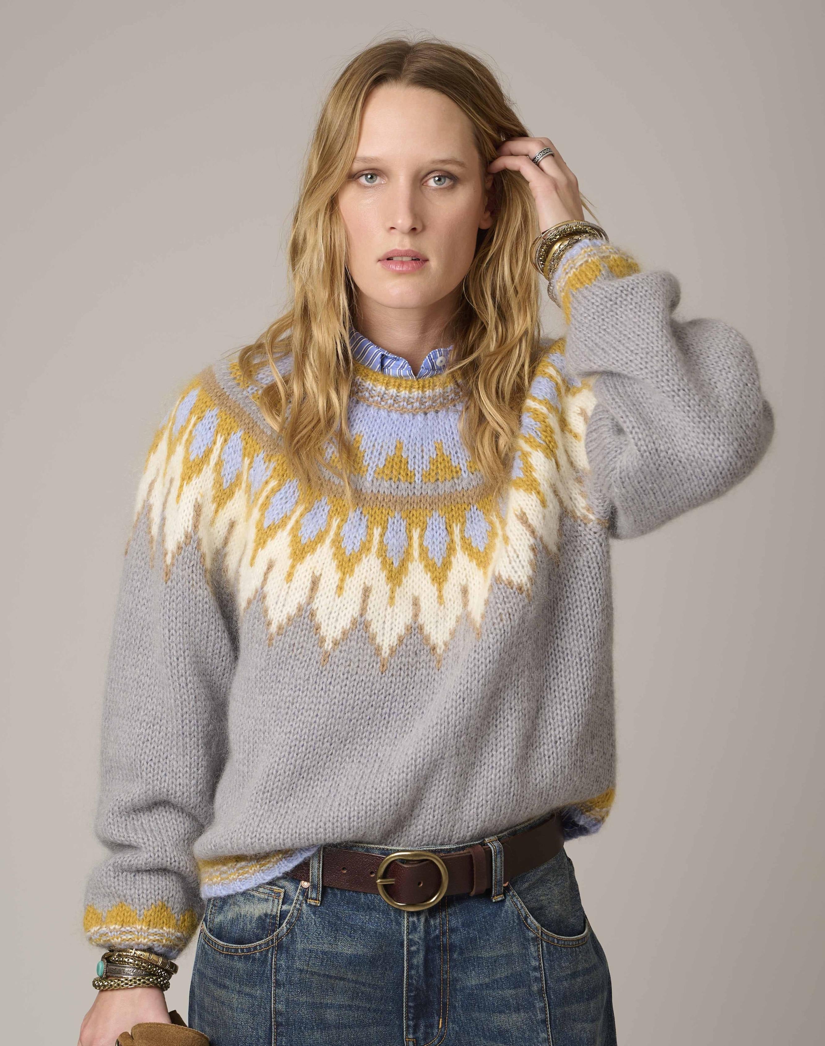 A woman modeling a gray knit sweater with a yellow and cream geometric yoke pattern, paired with blue jeans and a brown belt, against a solid light gray studio background.