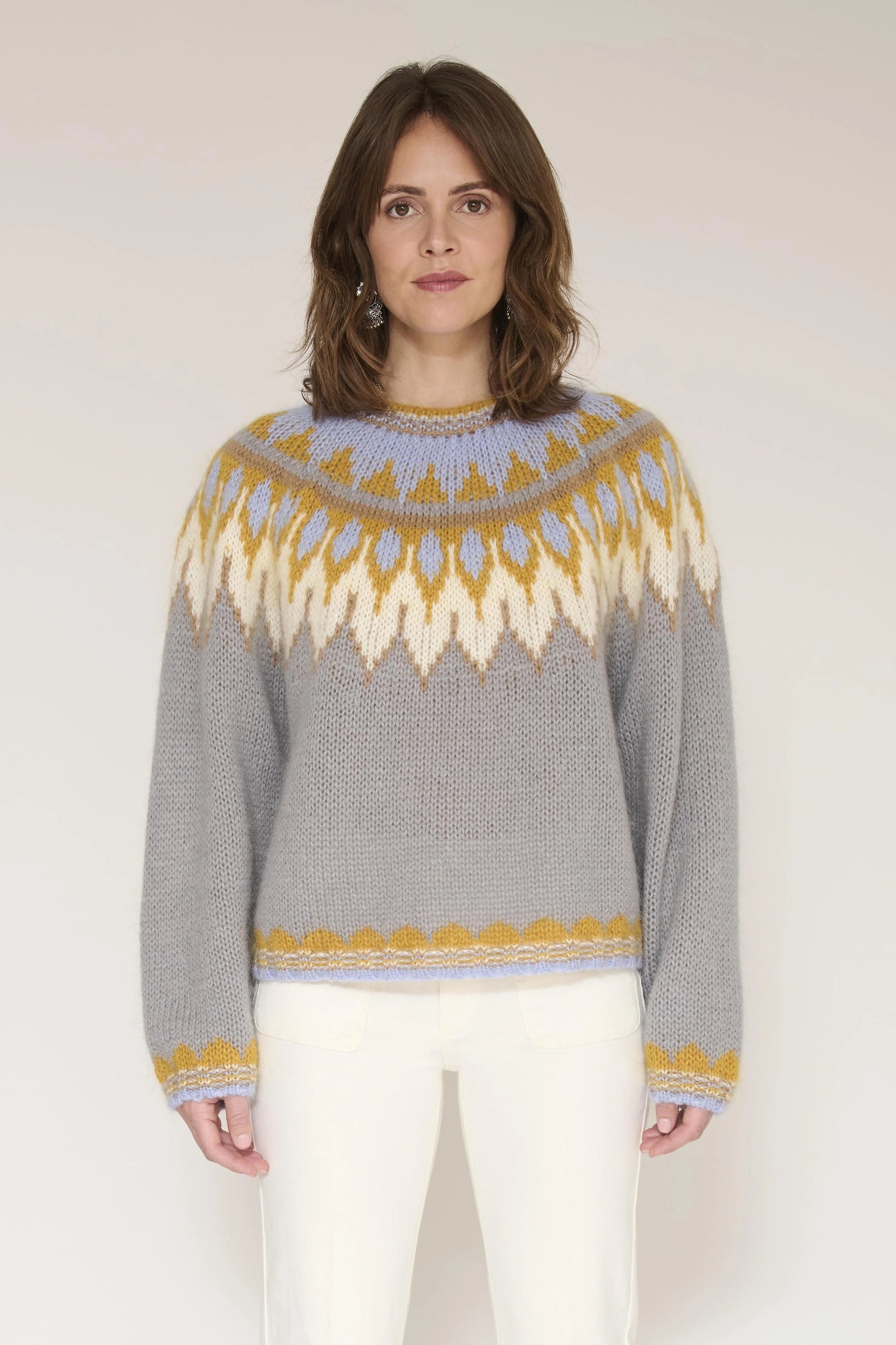 A woman wearing a gray knit sweater with a yellow and cream geometric pattern across the chest and collar, paired with white pants, standing against a light beige studio background.
