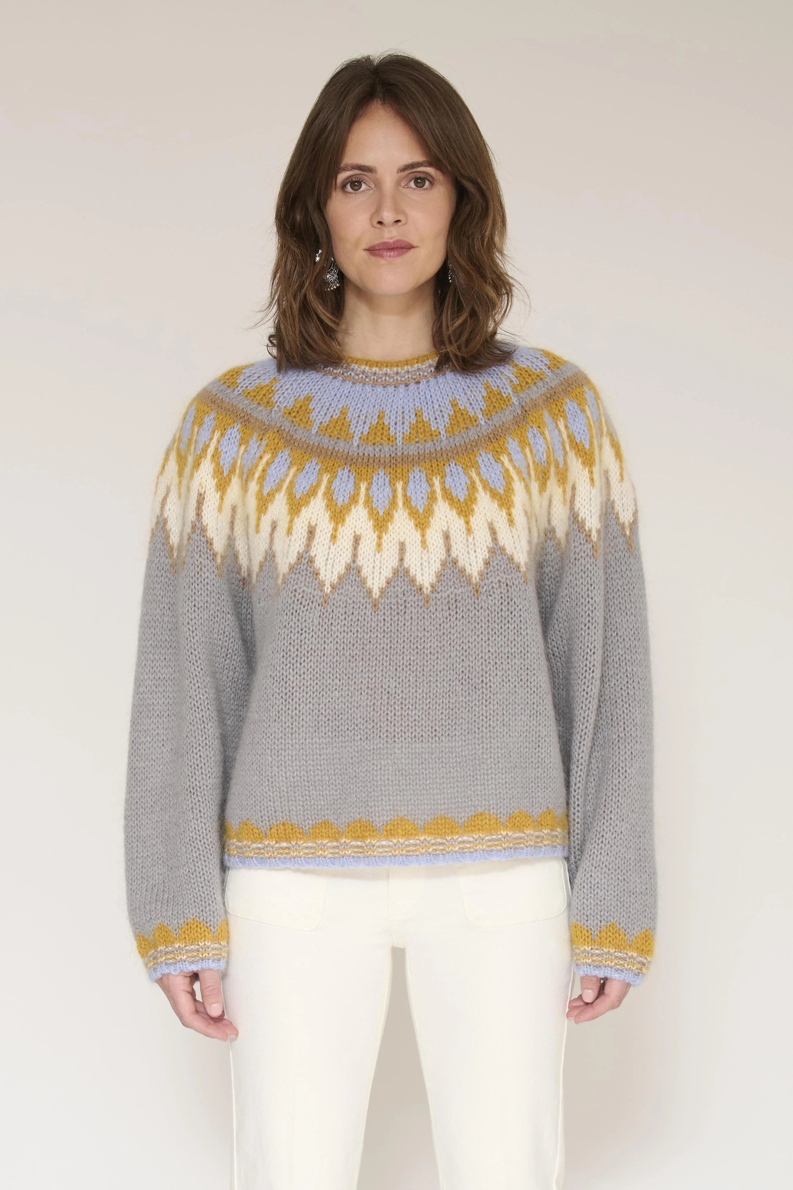 A woman wearing a gray knit sweater with a yellow and cream geometric pattern across the chest and collar, paired with white pants, standing against a light beige studio background.