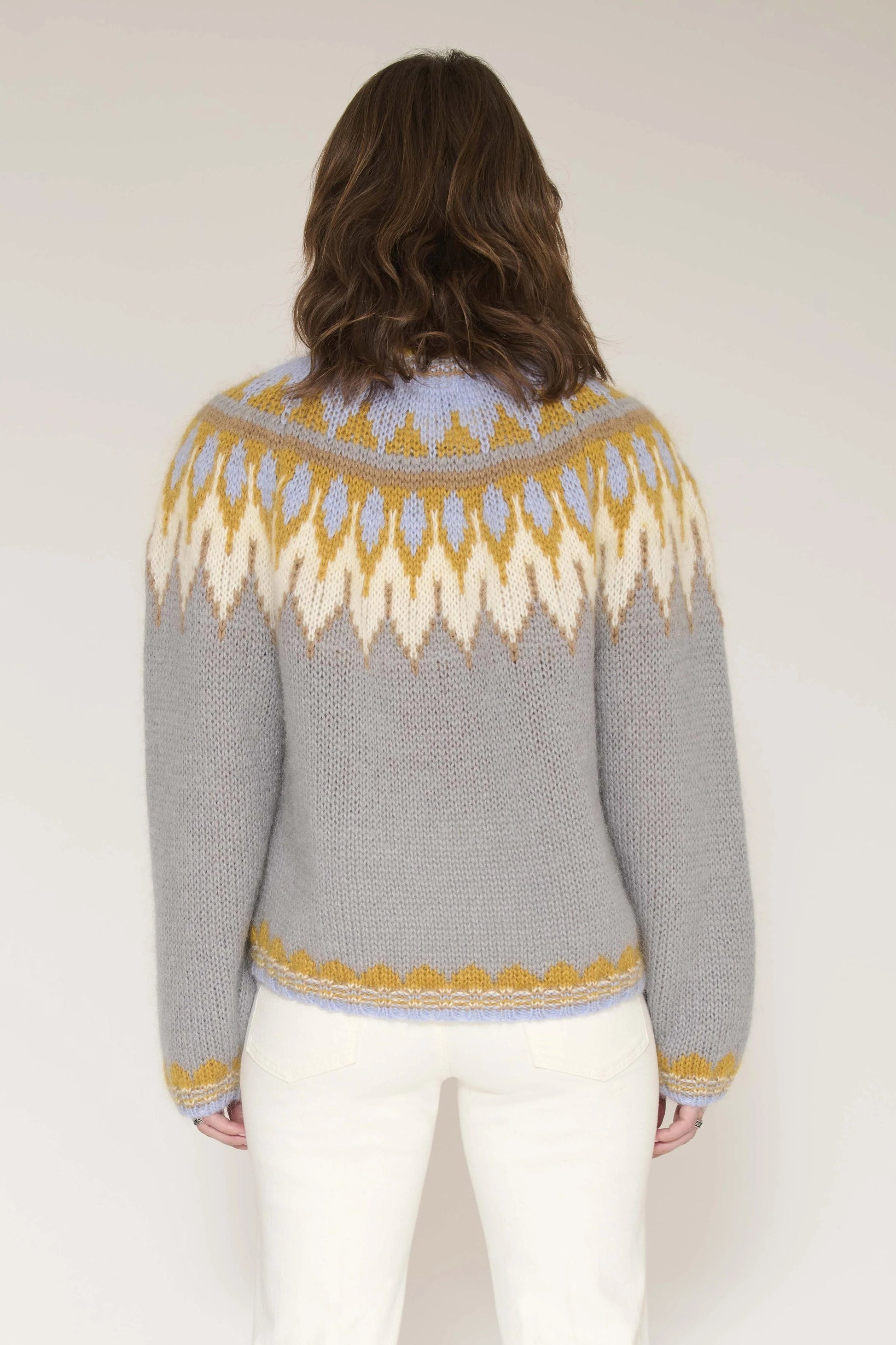 A woman viewed from behind wearing a gray knit sweater with a yellow and cream geometric pattern across the shoulders and upper back, paired with white pants, against a plain light background.