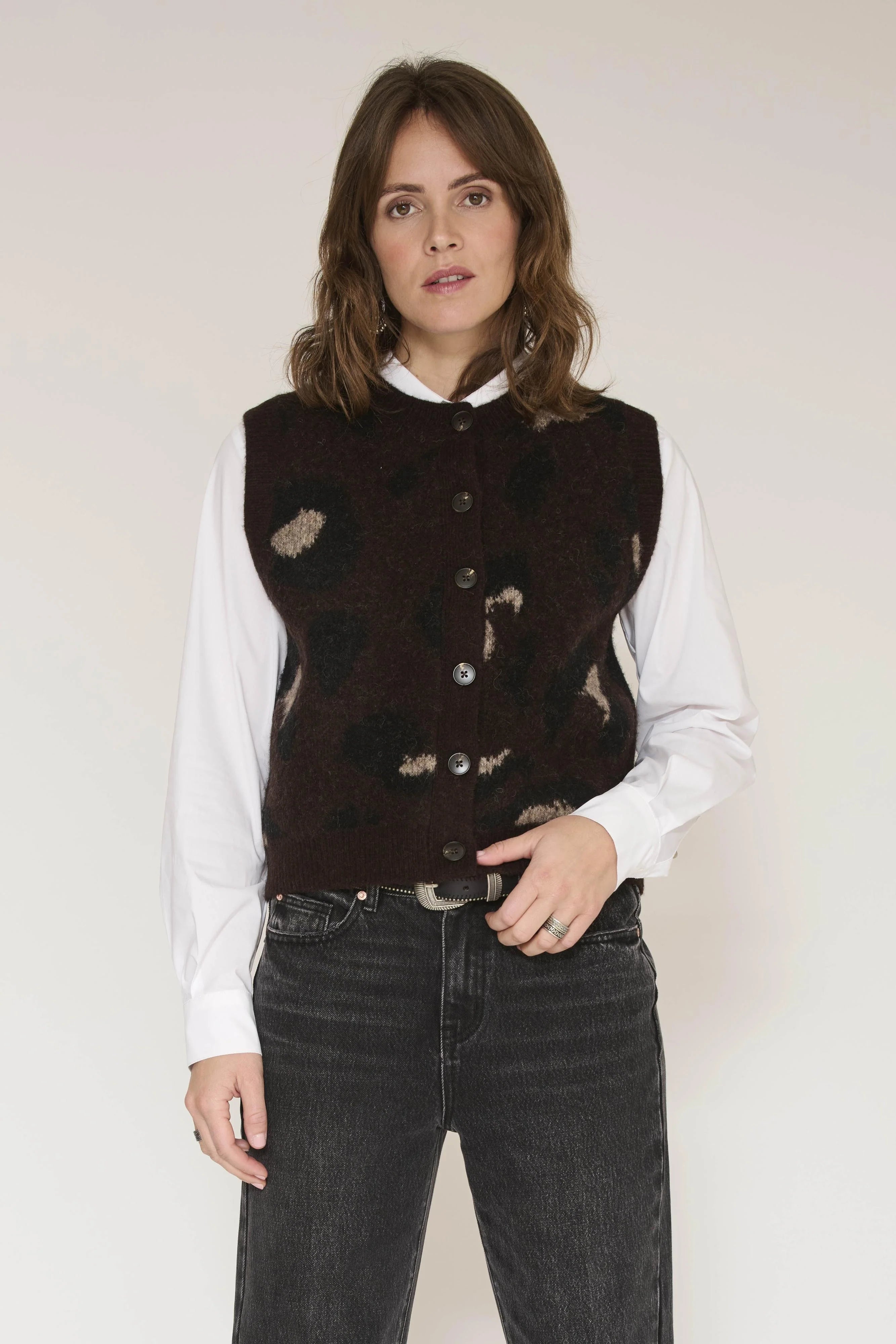 A woman modeling a dark brown patterned vest over a white collared shirt and dark gray jeans, standing against a plain light background.