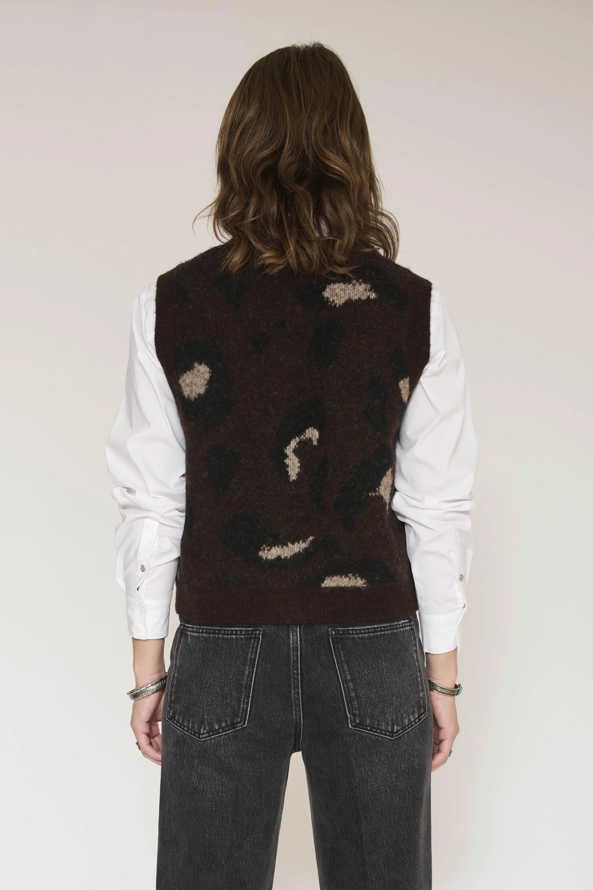A person stands facing away, wearing a white long-sleeve shirt, black jeans, and the MKTStudio Koba Knit Vest in brown layered over the shirt.