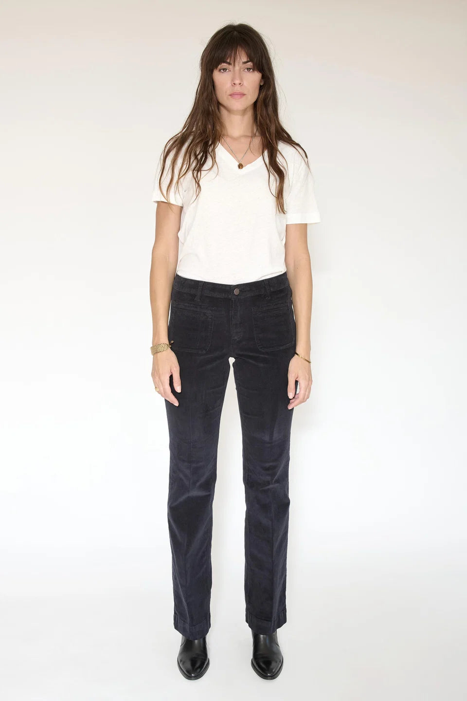 A woman stands against a plain background, wearing a white V-neck T-shirt, the Diana Velvet-Cord Trouser by MKTStudio with a flared silhouette in dark velvet, black boots, and minimal jewelry.