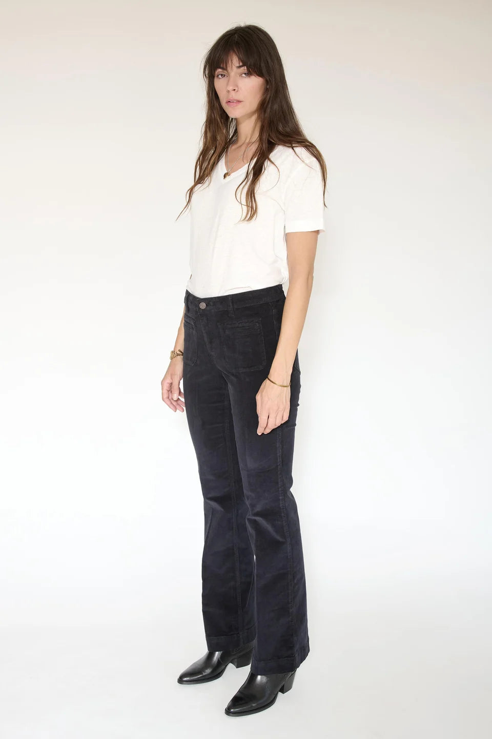 A woman stands against a plain background wearing a white t-shirt, black heeled boots, and the Diana Velvet-Cord Trouser by MKTStudio—dark corduroy pants featuring a subtly flared silhouette.