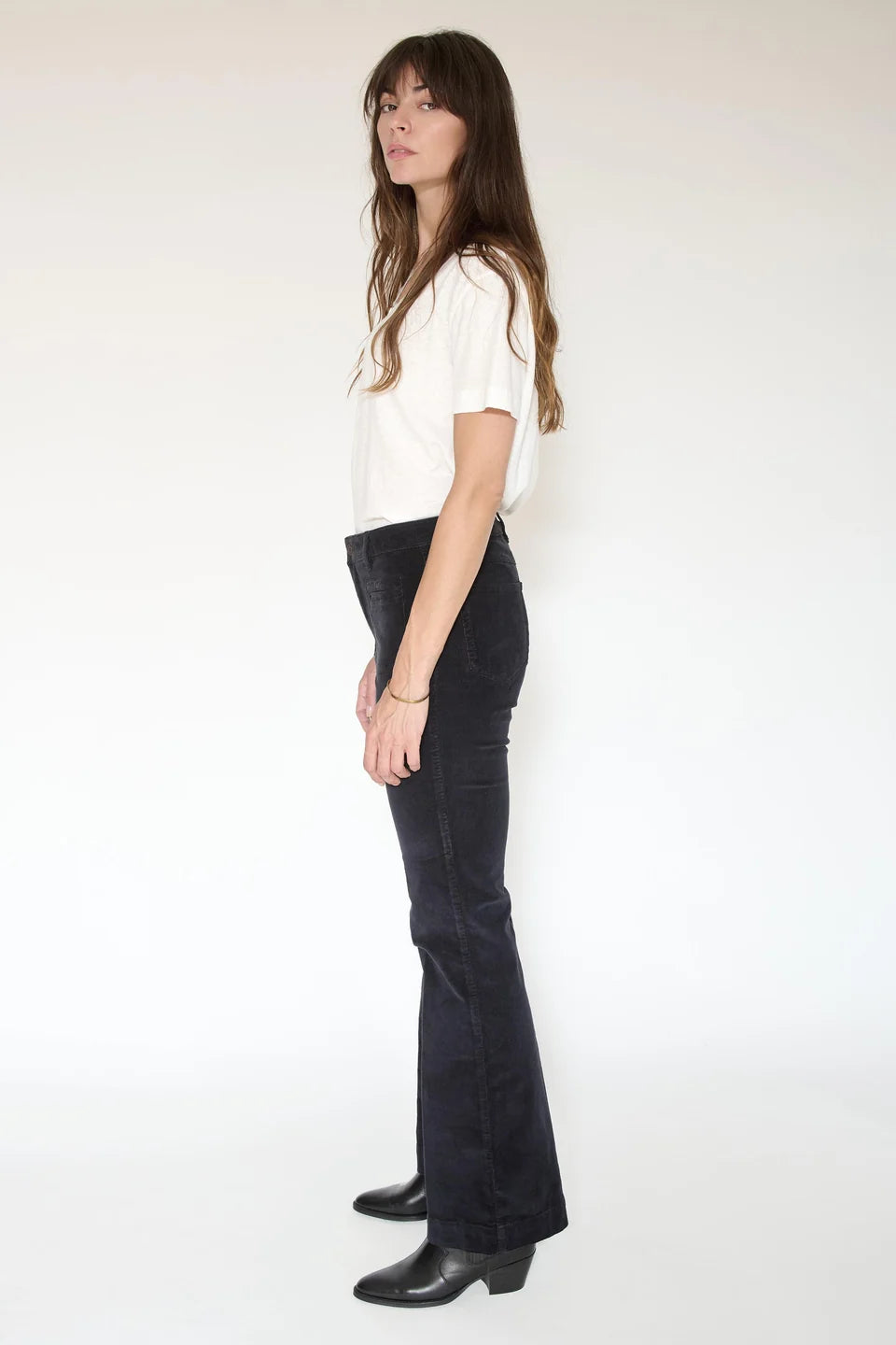 A woman stands in profile against a plain background, wearing a white t-shirt, black heeled boots, and MKTStudio’s The Diana Velvet-Cord Trouser.