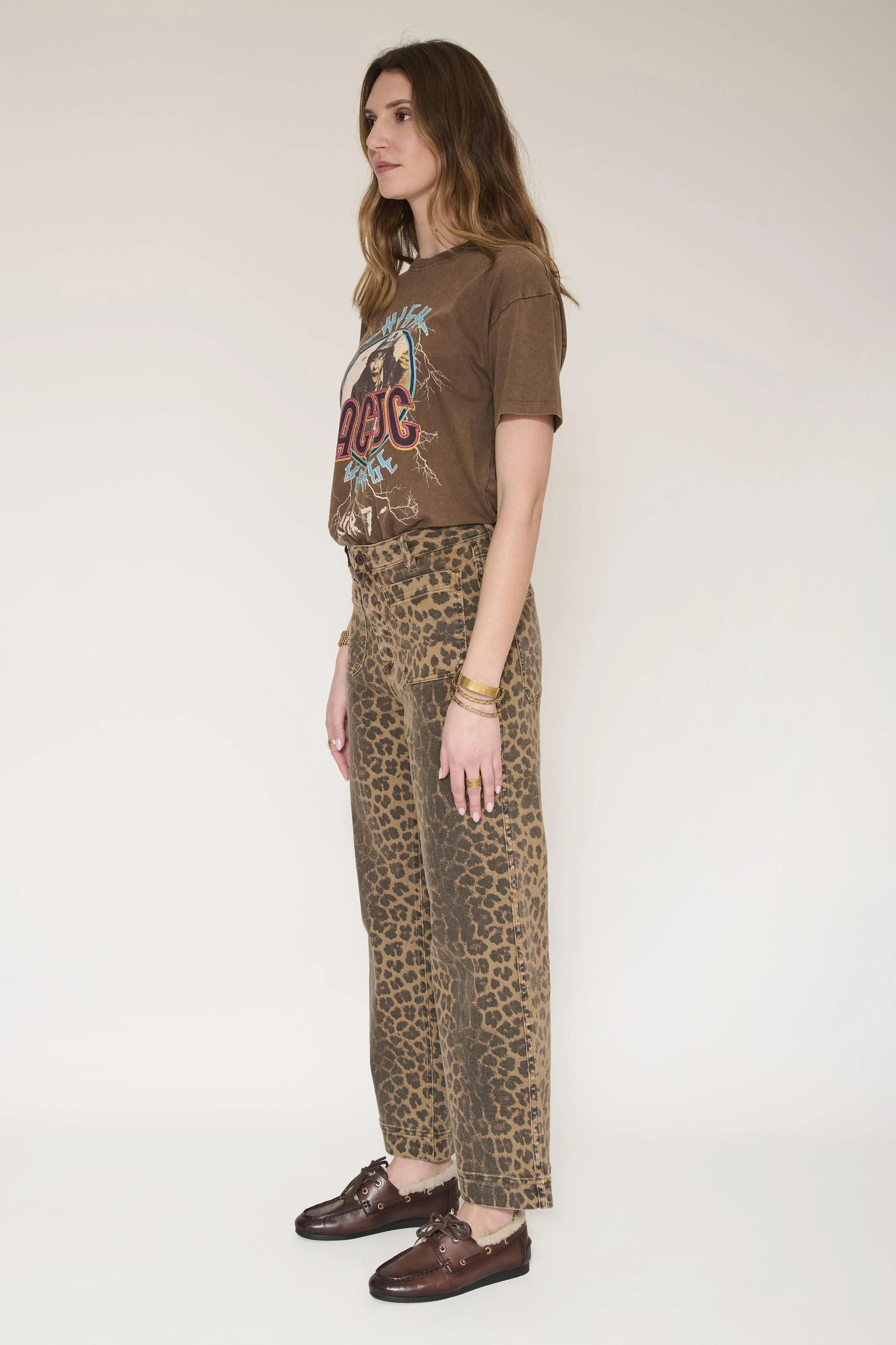 A woman stands in profile wearing a brown graphic T-shirt, leopard-print wide-leg pants, and brown leather loafers against a plain off-white studio background.