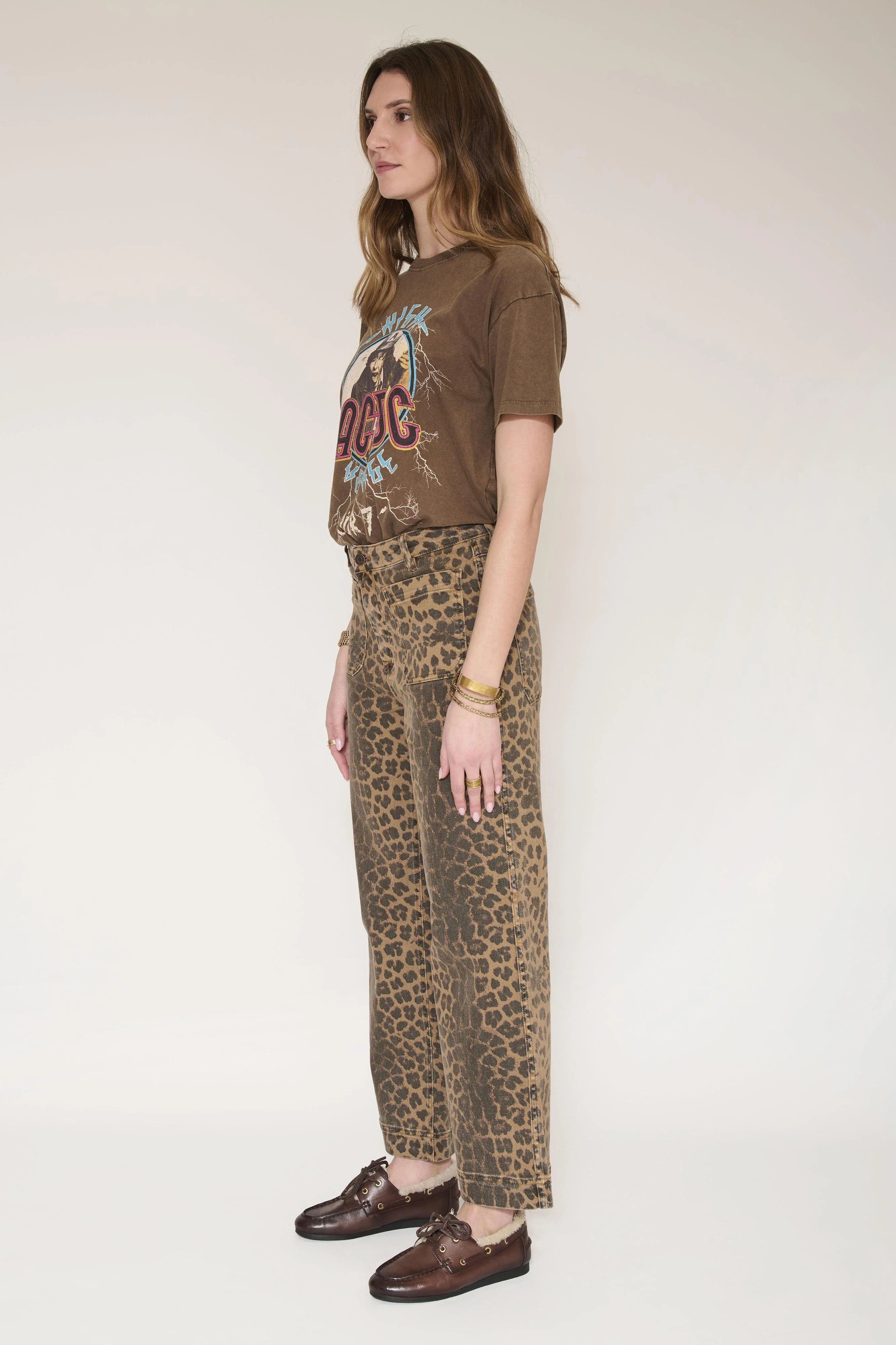 A woman stands in profile wearing a brown graphic T-shirt, leopard-print wide-leg pants, and brown leather loafers against a plain off-white studio background.
