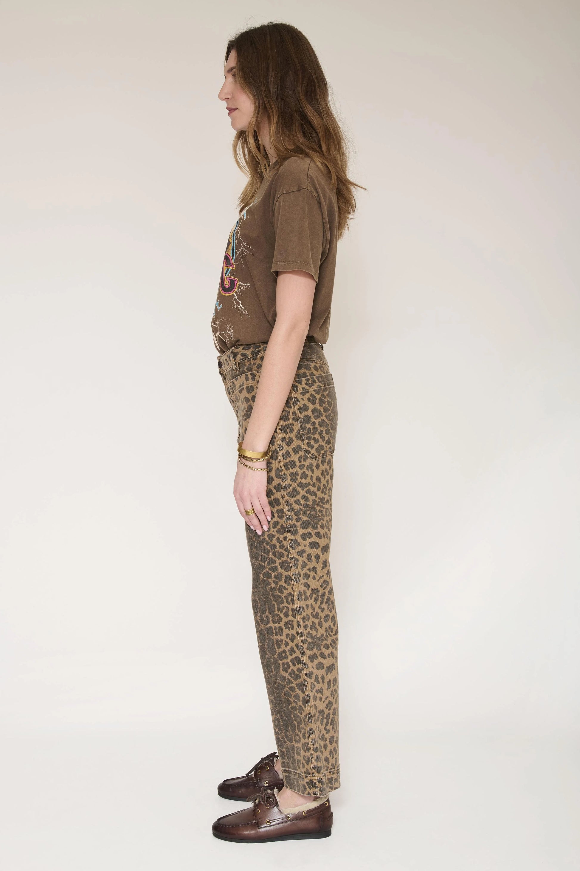 A woman shown in profile wearing a brown graphic t-shirt, leopard-print trousers, and dark brown leather loafers against a plain off-white studio background.