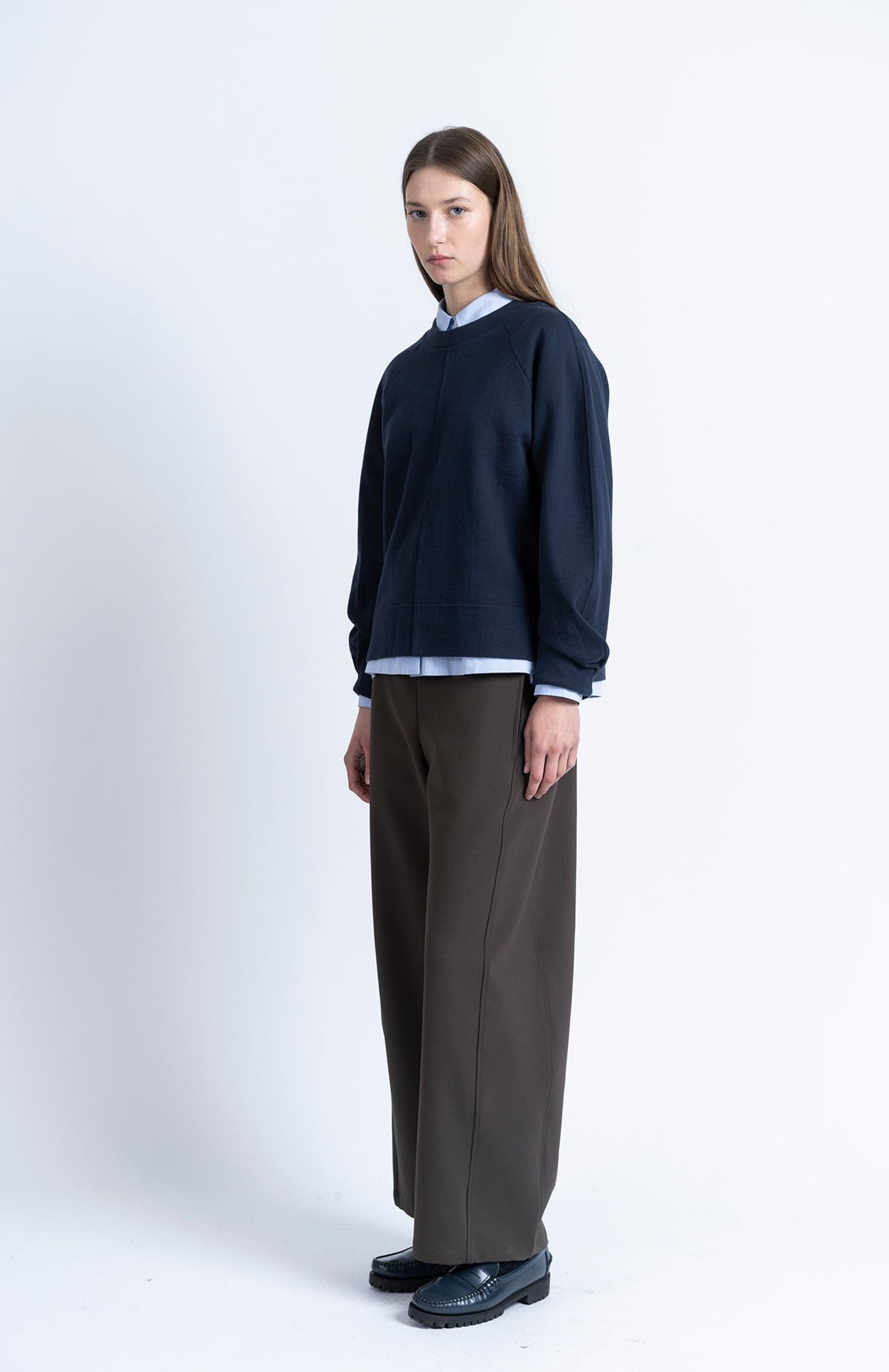 A person wearing a navy Loreak Mendian Celine Sweatshirt over a light blue collared shirt, paired with dark wide-leg trousers and dark loafers.