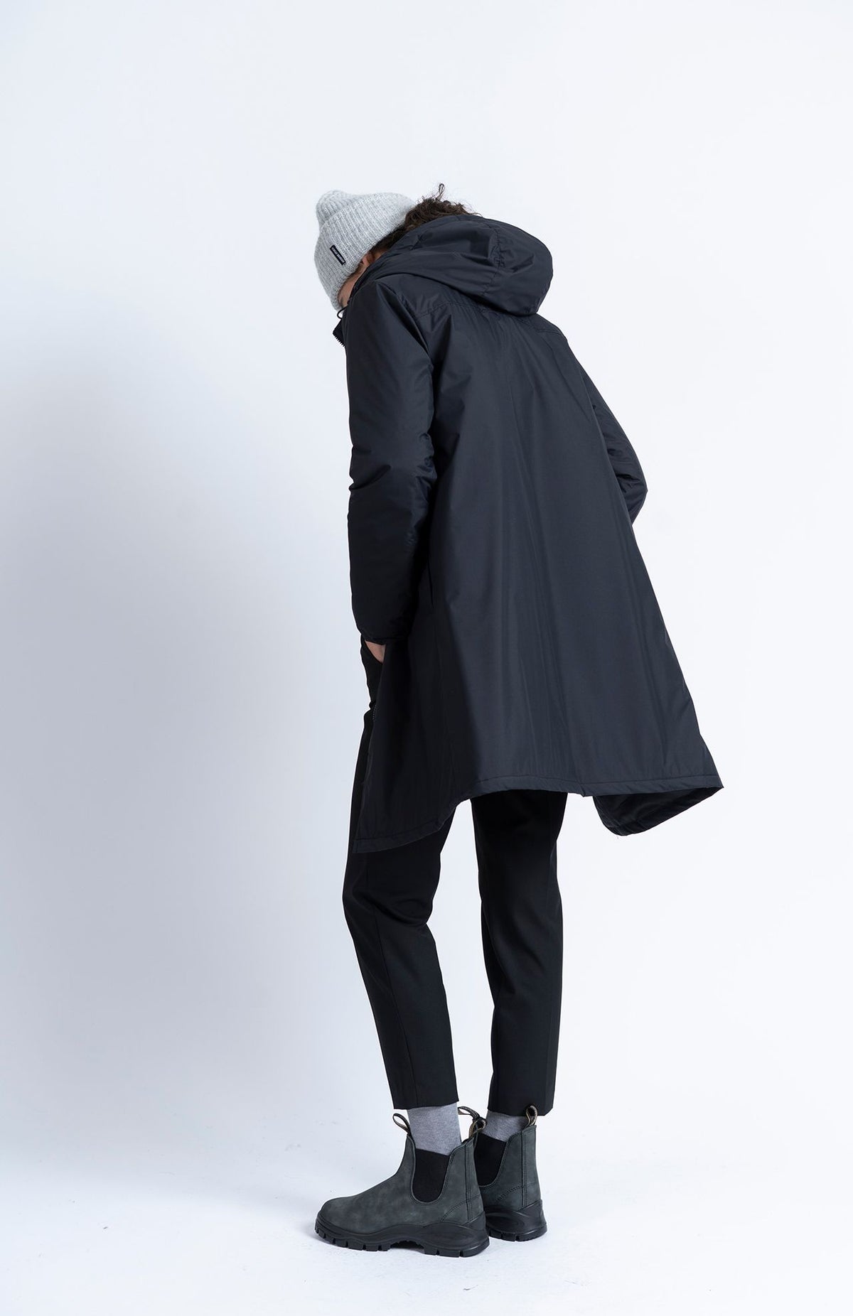 Person seen from behind wearing a black hooded parka, Alaiak Trouser - Black joggers, gray beanie, and black boots.